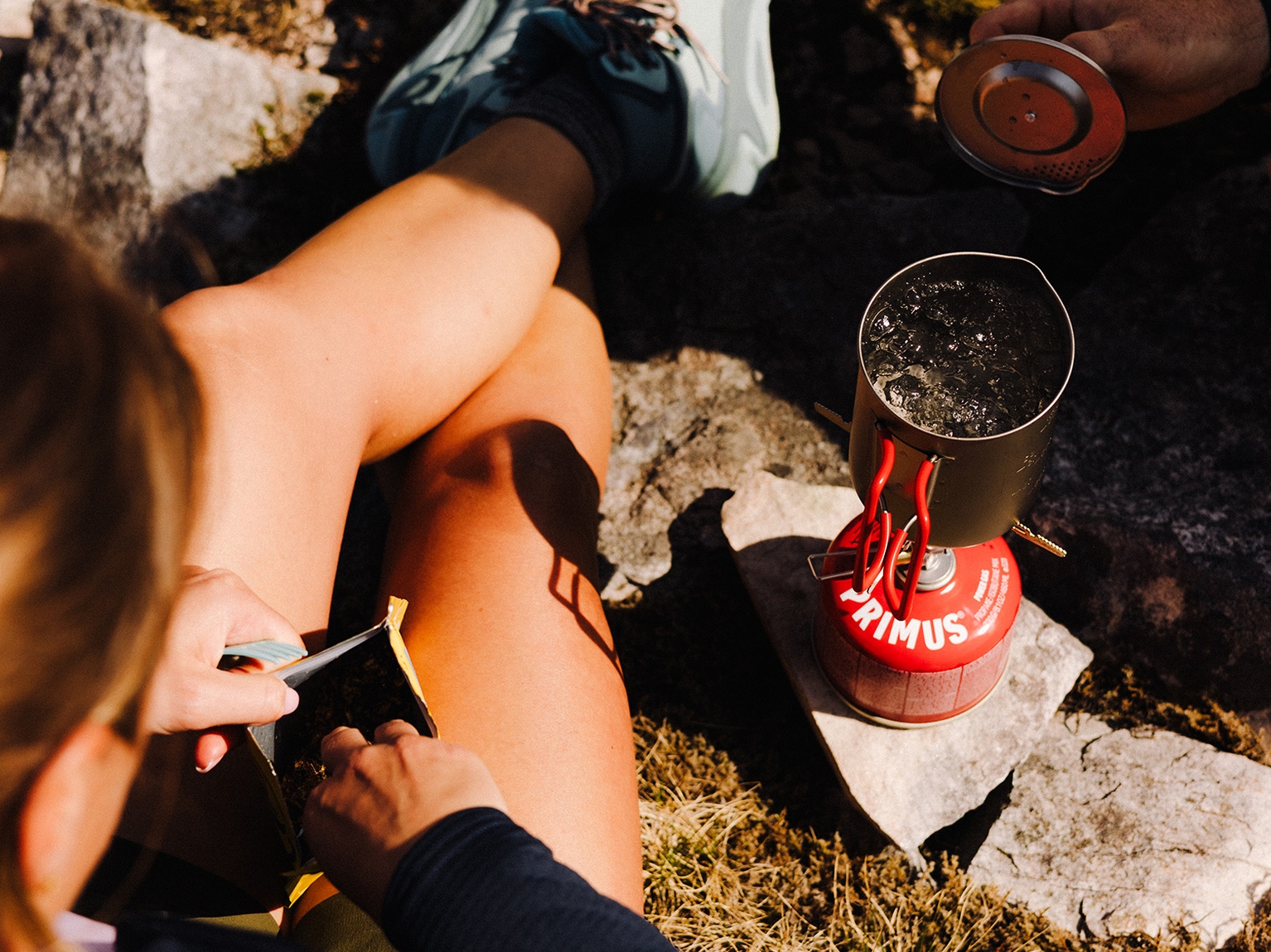 Person camping outdoors using a portable stove to boil water, with a food packet being opened nearby on rocky ground.