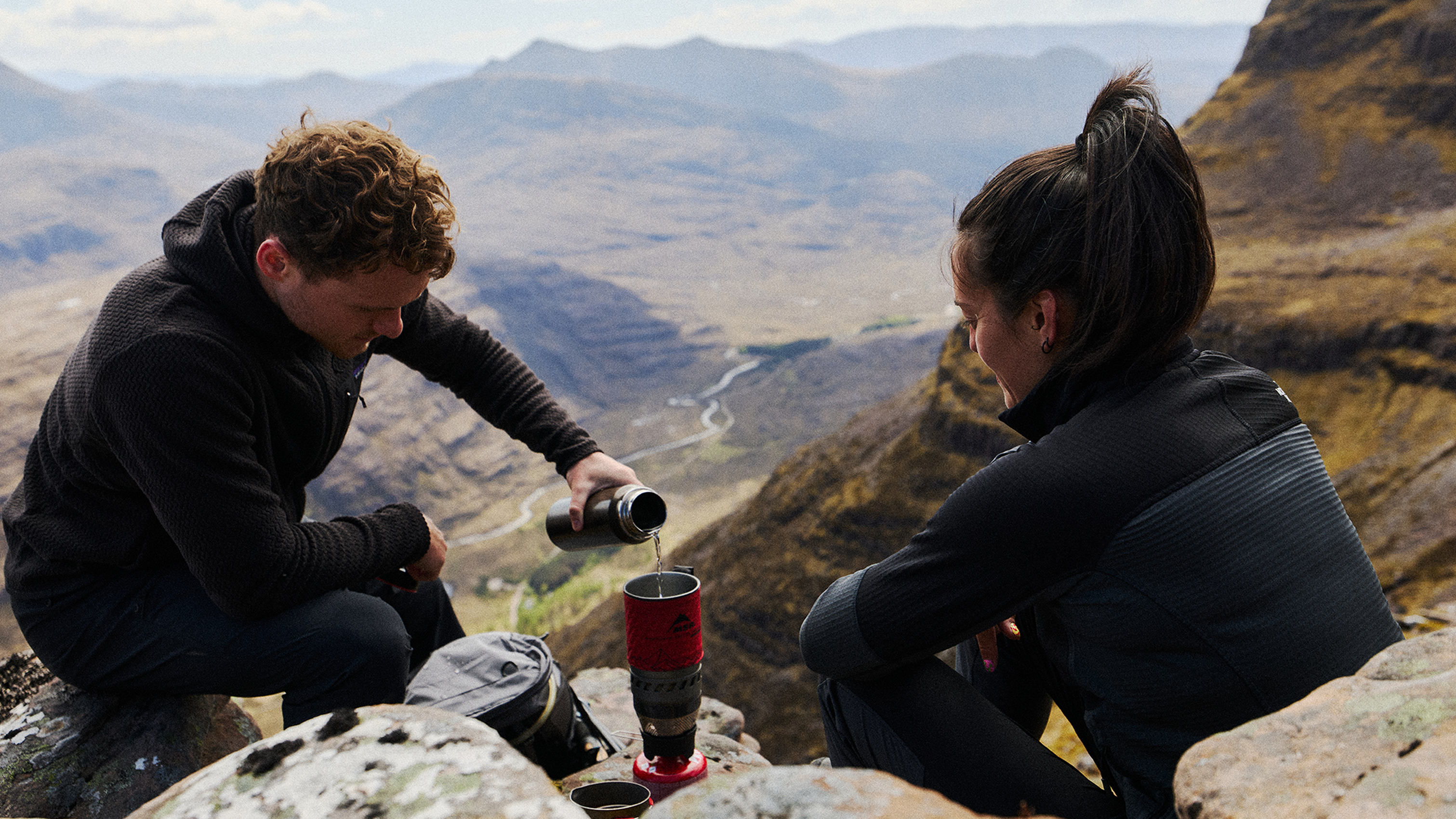Two people enjoy a break on a mountain, with one pouring a drink. Expansive valley and rugged terrain stretch in the background.