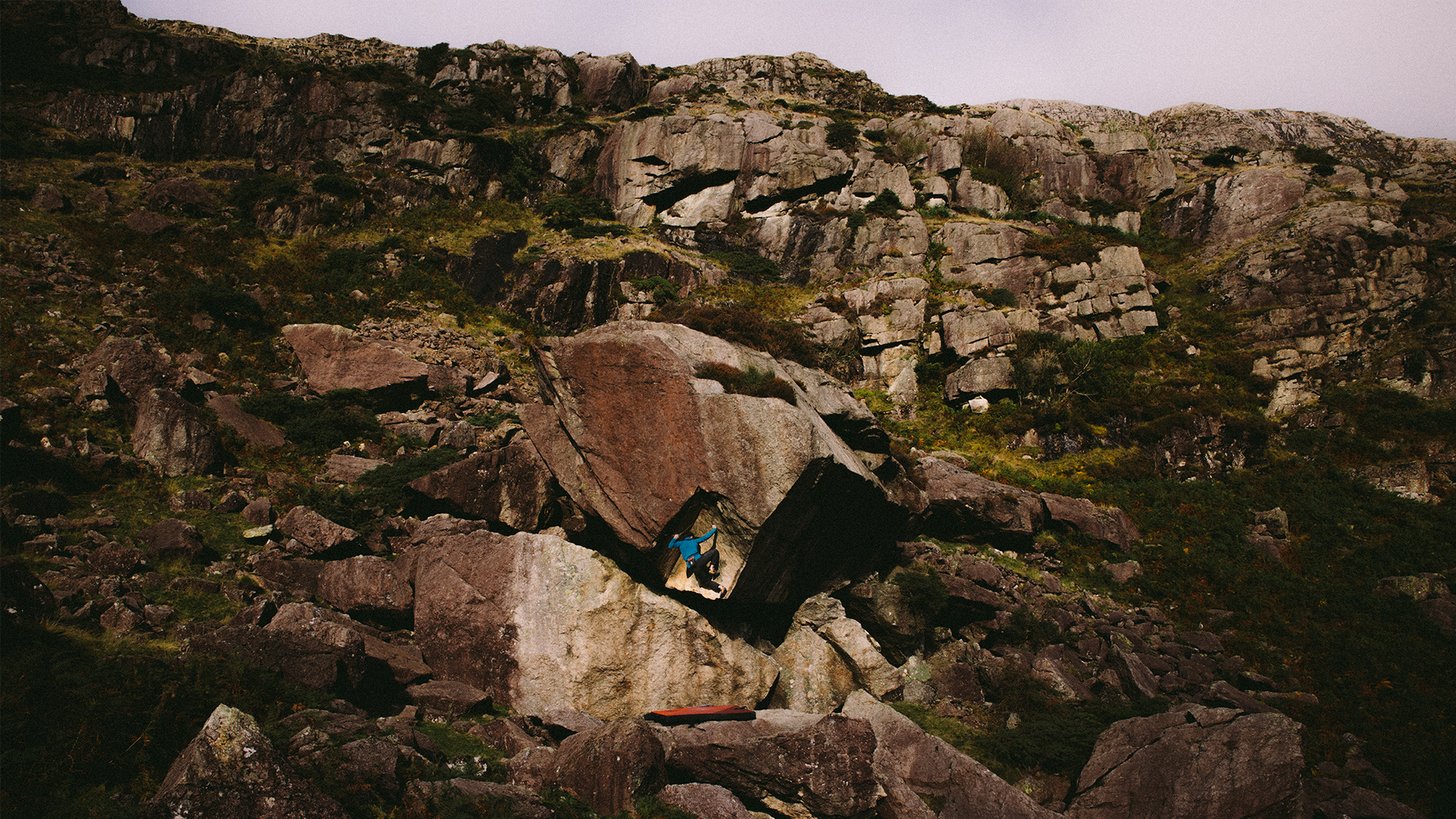 Climber in a blue jacket navigating a large, rugged rock formation on a steep, rocky hillside under a cloudy sky.
