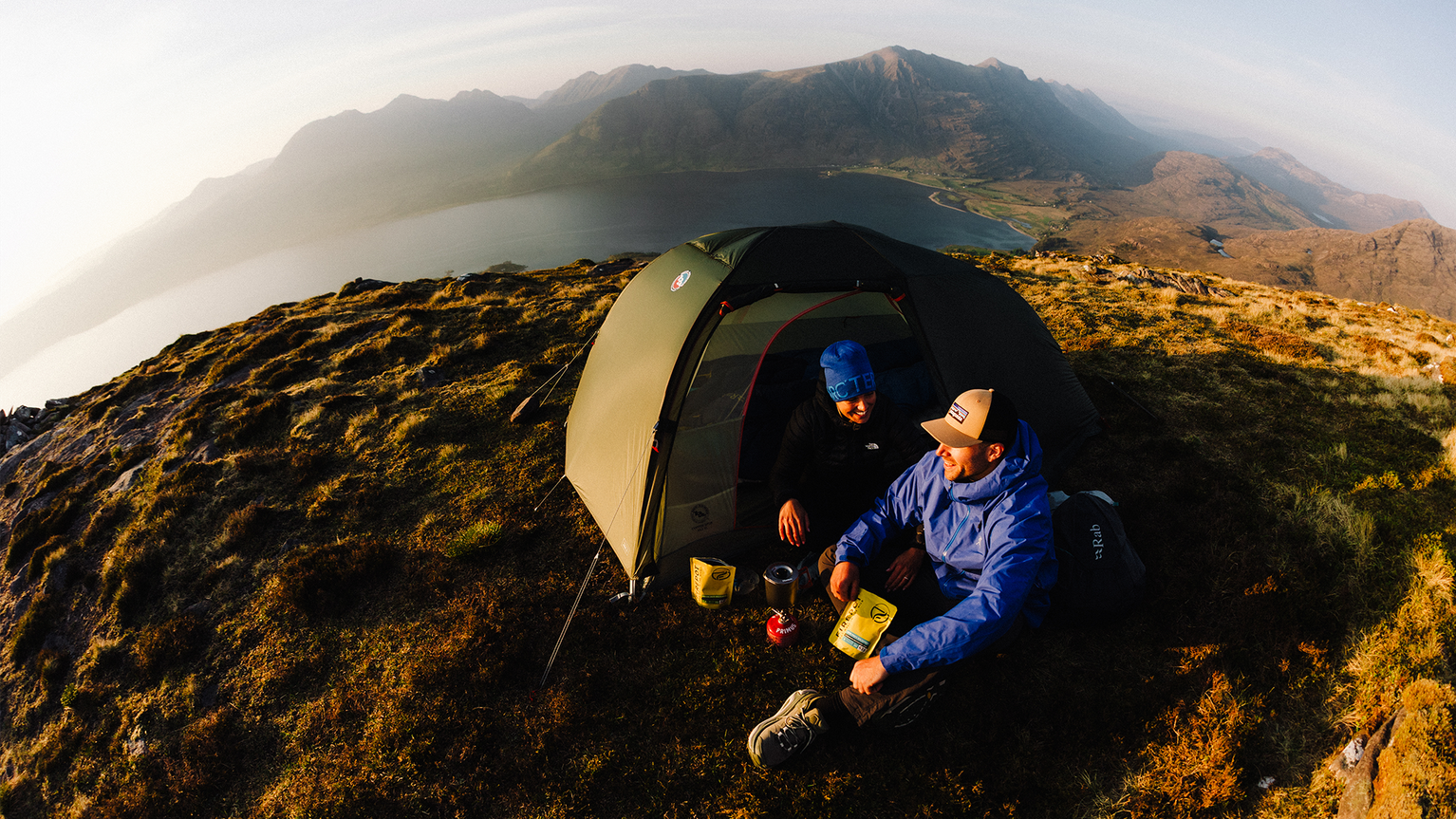 Two people in a tent camping on a mountain, eating from packets. Scenic view of a lake and mountains in the background.