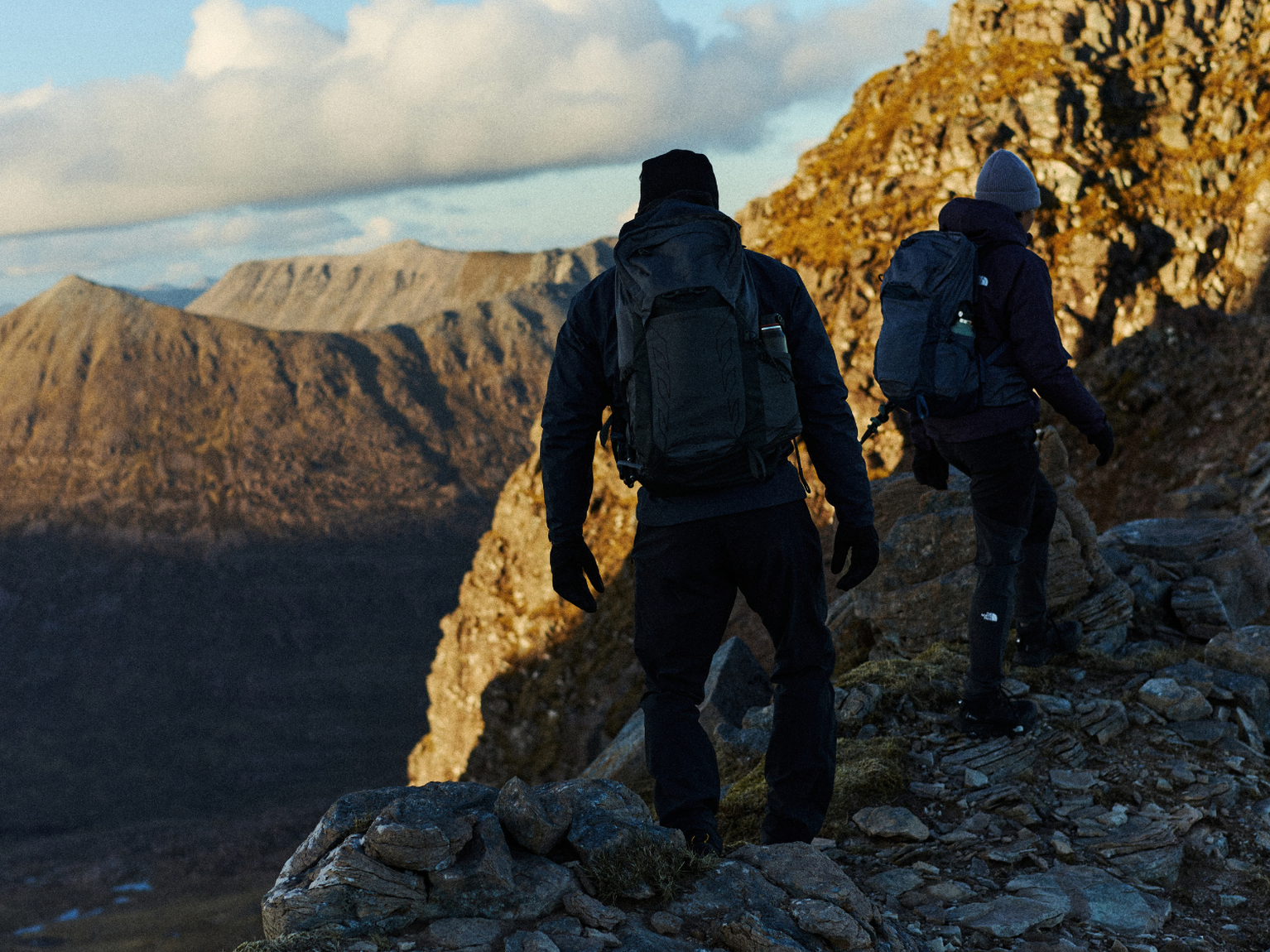 Two hikers with backpacks traverse a rocky mountain trail, with expansive views of rugged peaks and cloudy skies in the background.