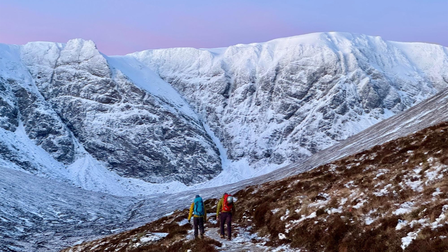 Two hikers in vibrant jackets trek towards a snow-covered mountain range under a pastel sky, navigating a rugged trail.