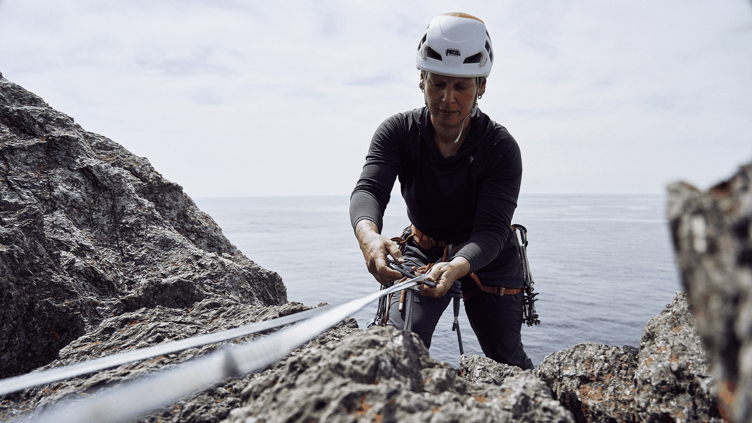 Climber wearing a helmet and harness secures a rope on a rocky cliff near the sea under a clear sky.