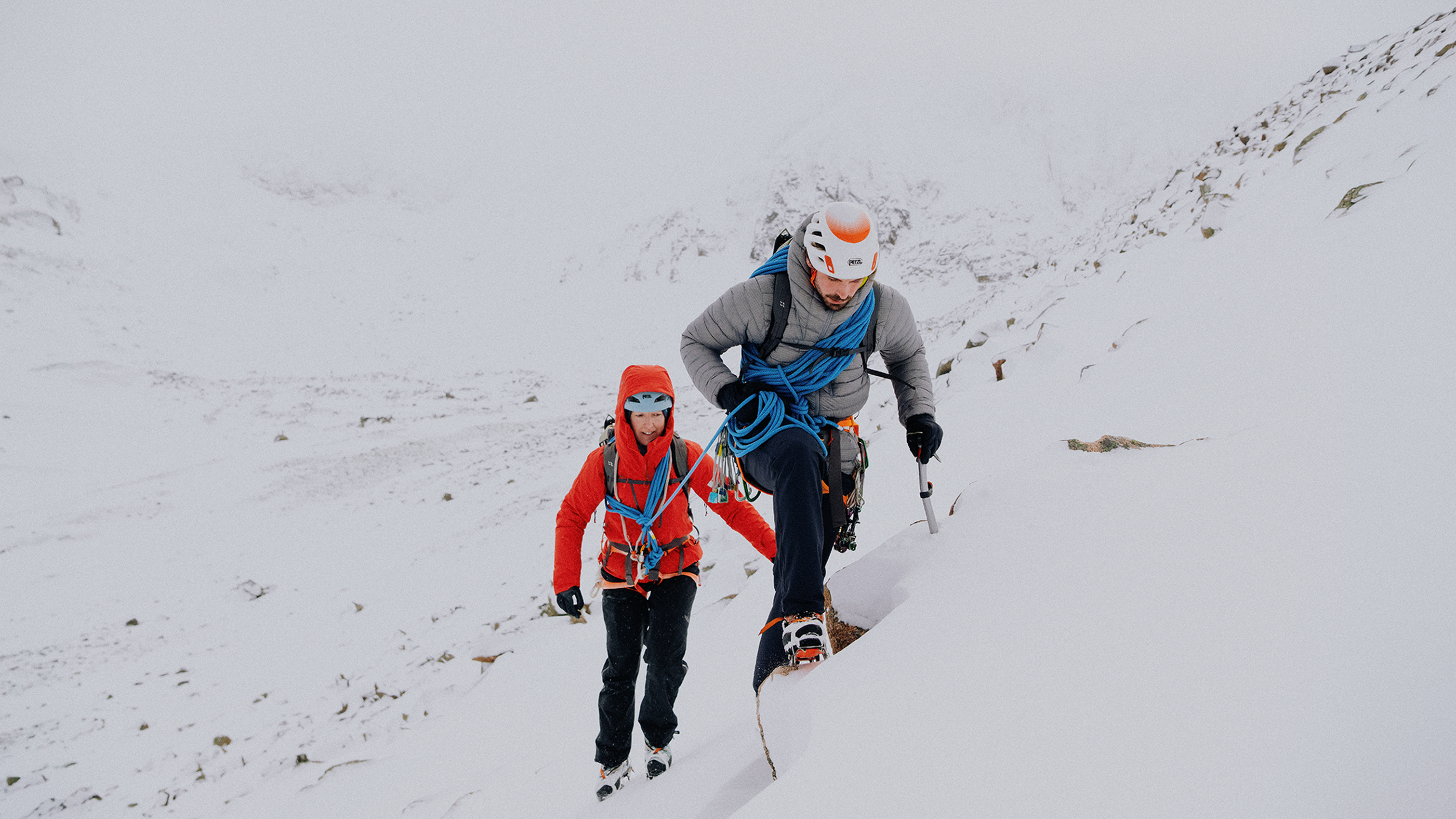Two climbers, equipped with ice axes and ropes, ascend a snowy mountain slope in challenging conditions, demonstrating expertise and resilience.