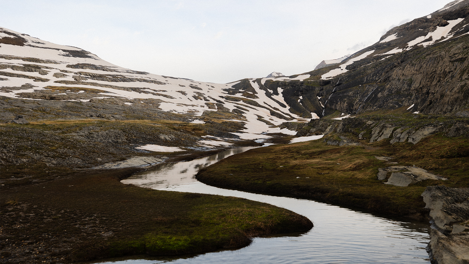 A winding stream flows through a mountainous landscape with patches of snow and rocky terrain under a clear sky.