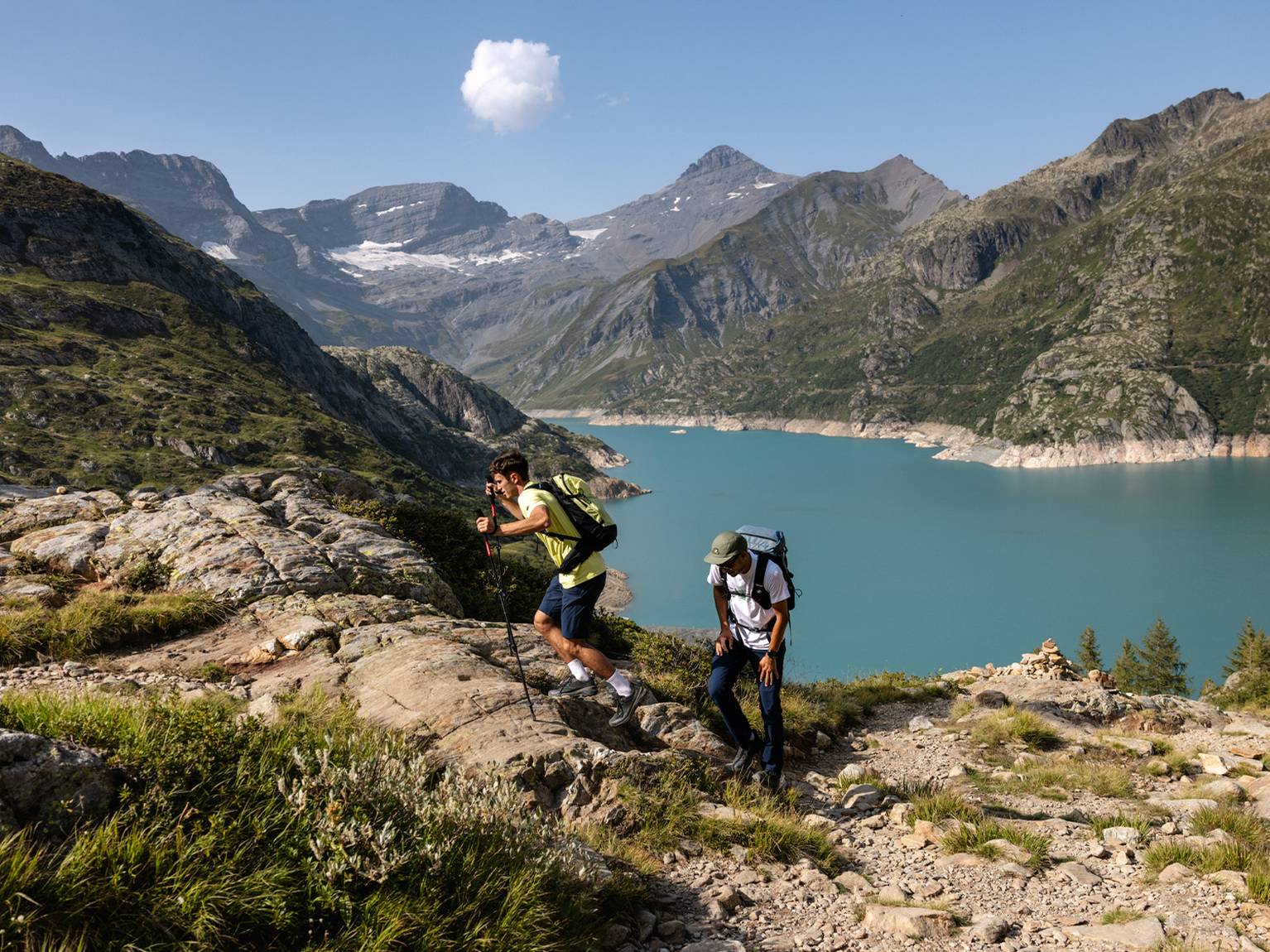 Two hikers climbing a rocky trail with trekking poles, overlooking a blue lake and surrounded by mountain peaks and lush greenery under a clear sky.
