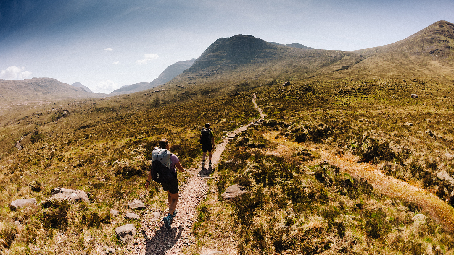 Two hikers walking along a narrow trail surrounded by rugged grassy hills under a clear blue sky.