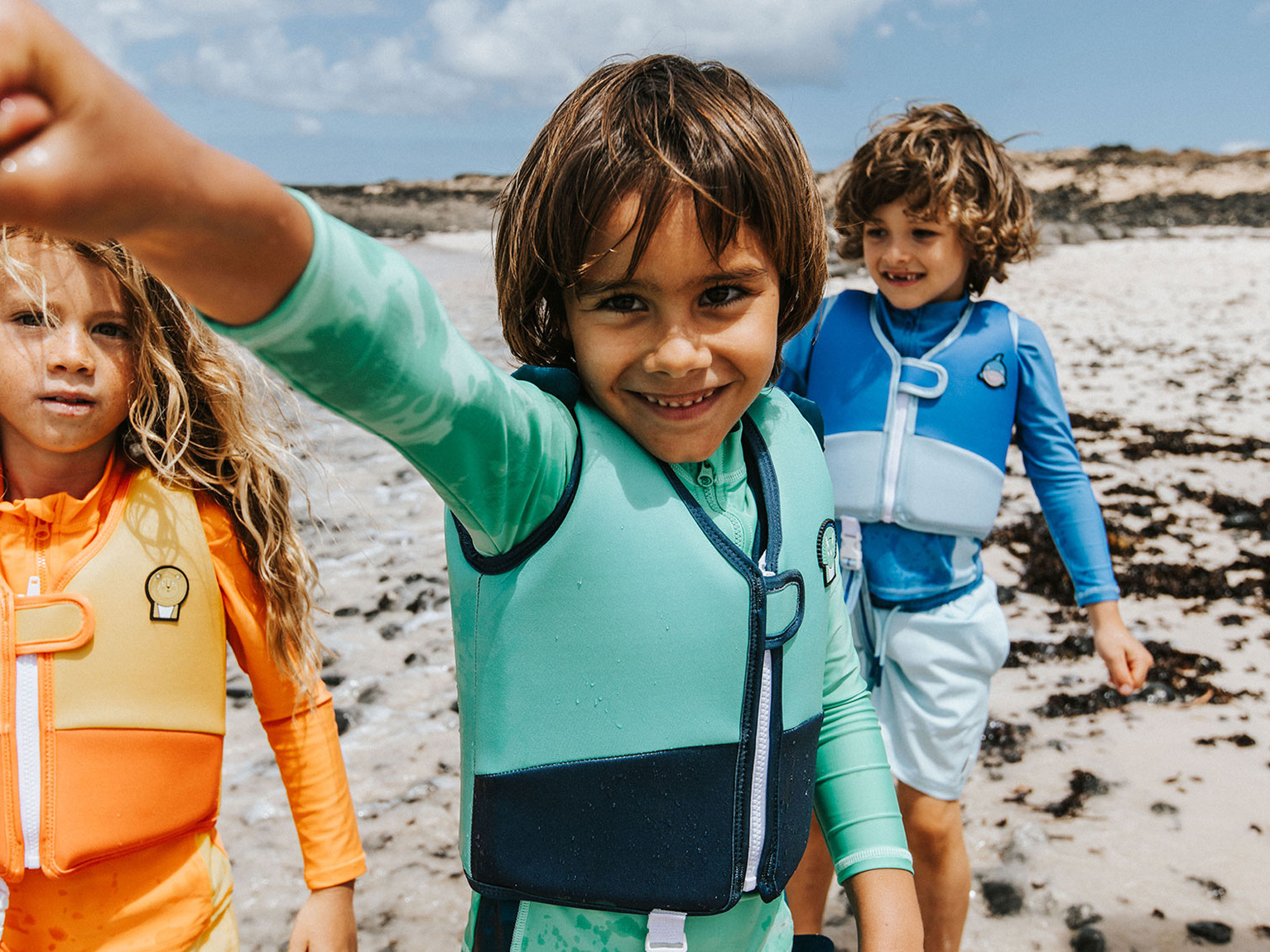 Three children in colourful wetsuits and buoyancy aids on a rocky beach, smiling and reaching towards the camera.
