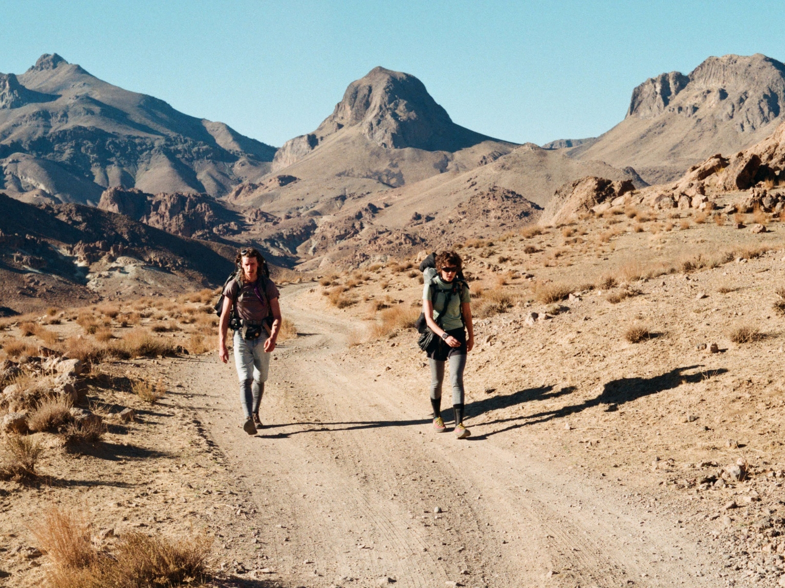 Two hikers walk along a dirt path in a mountainous landscape, surrounded by rugged terrain and clear blue skies.