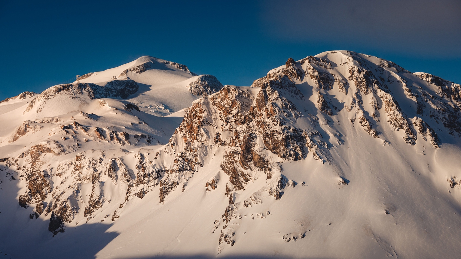 Snow-covered mountain peaks under a clear blue sky, with sunlight casting shadows on the rugged terrain.