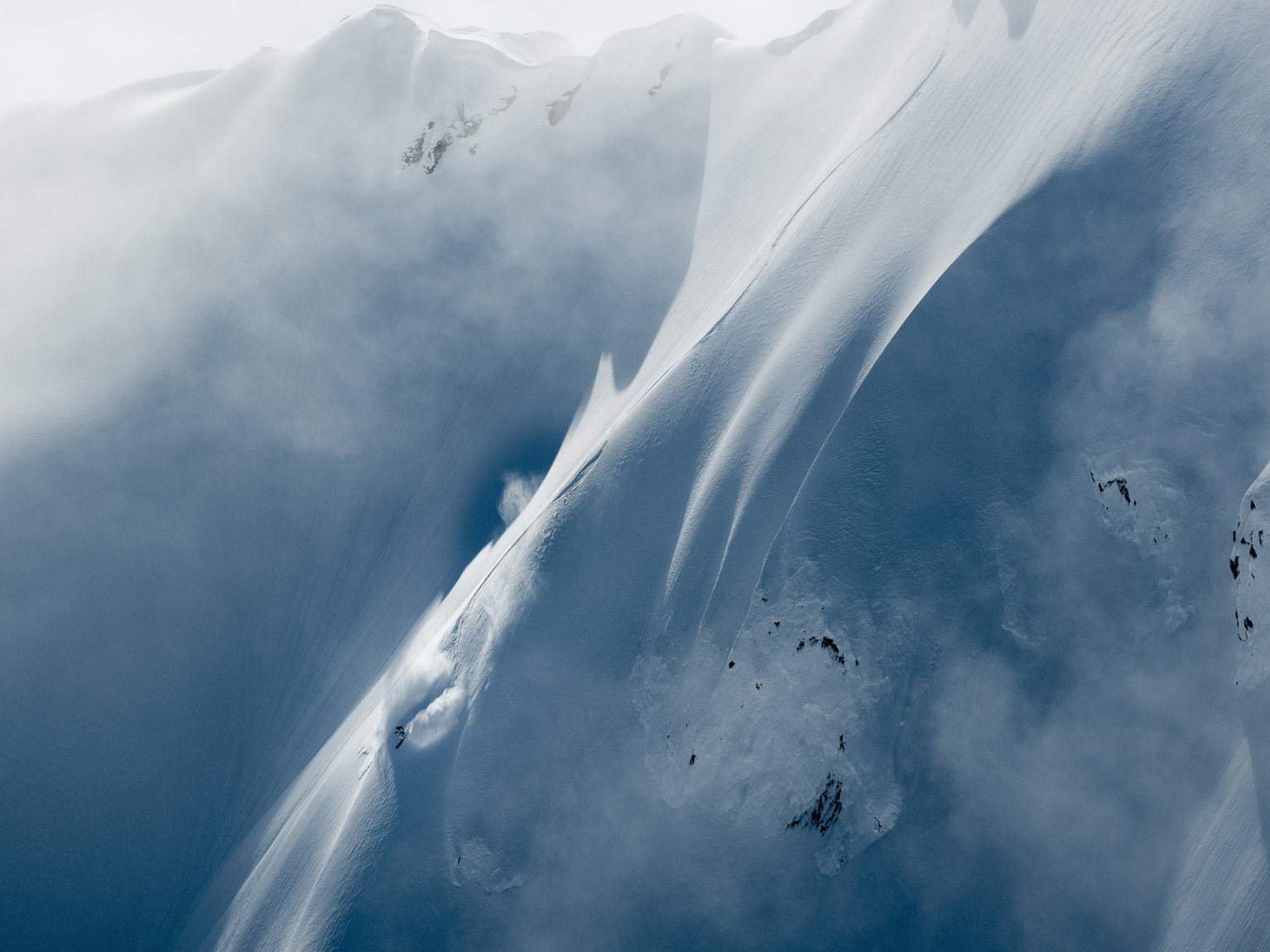 A lone skier gracefully descends a steep, snow-covered mountain slope under a cloudy sky.