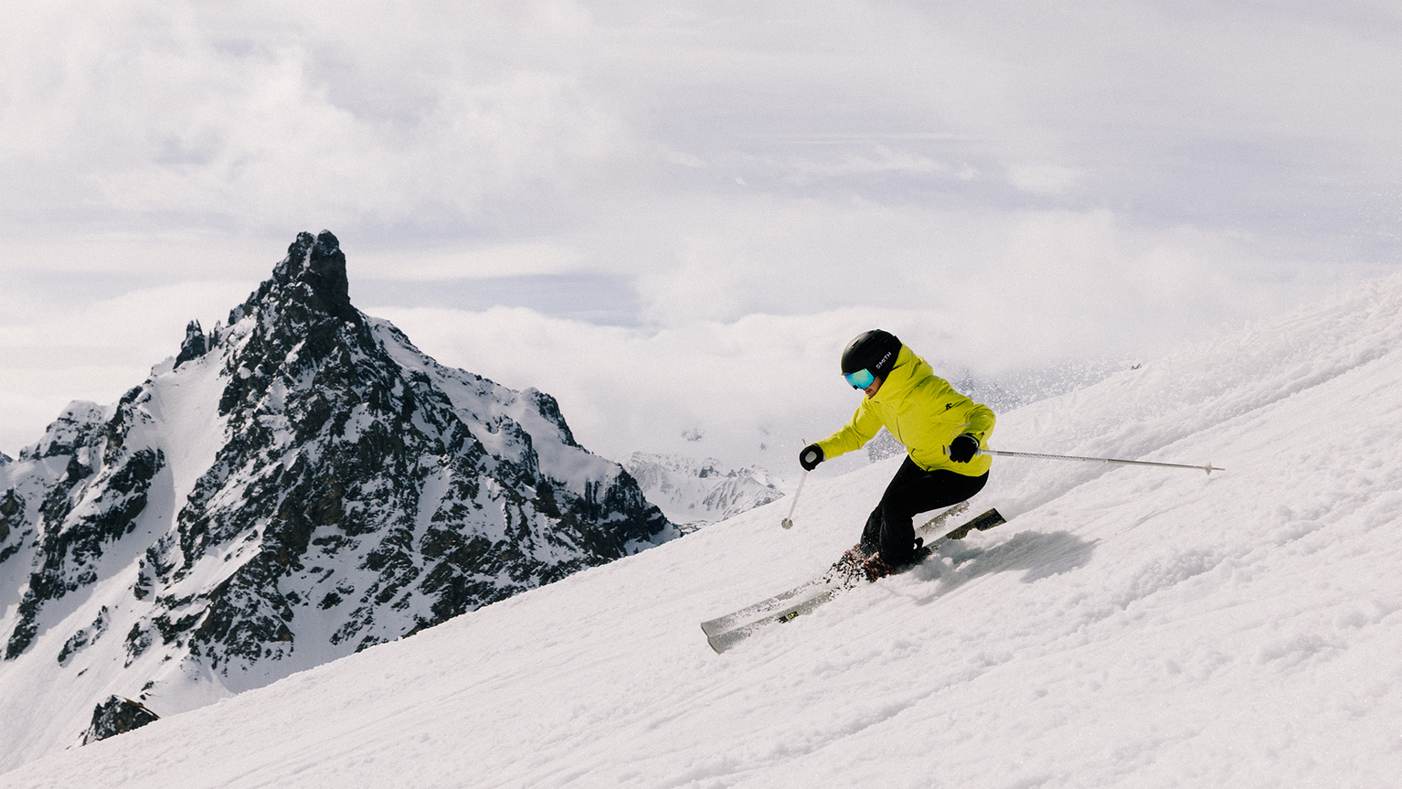 A skier in a bright yellow jacket descends a snowy slope with a jagged mountain peak in the background under a cloudy sky.