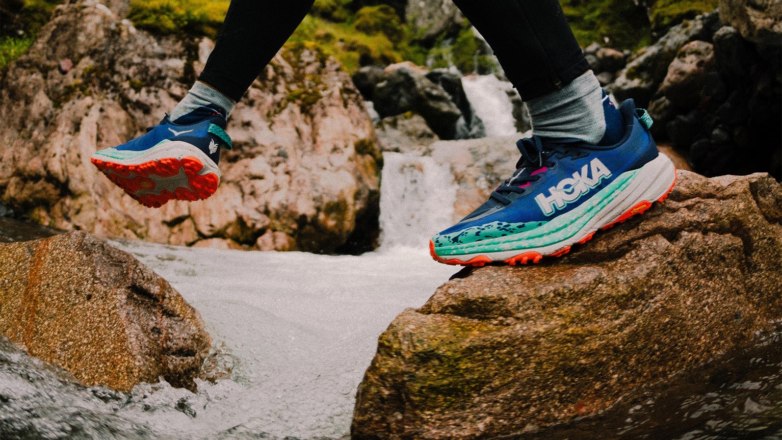 Close-up of a person wearing blue and orange Hoka trail running shoes, stepping on rocks across a flowing stream with a waterfall in the background.