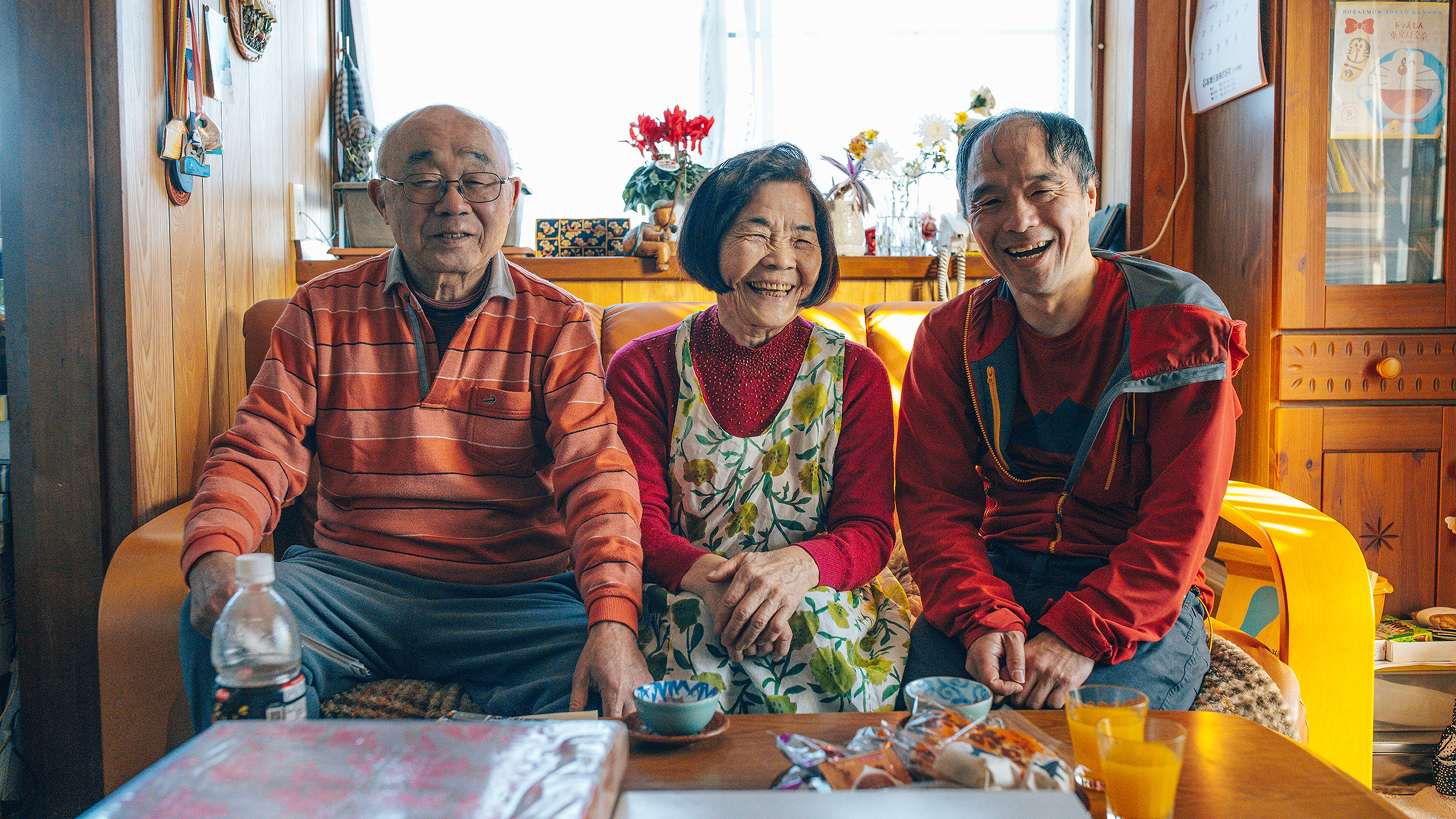Three people sitting on a sofa, smiling warmly in a cosy, well-lit room with wooden walls and colourful decorations.