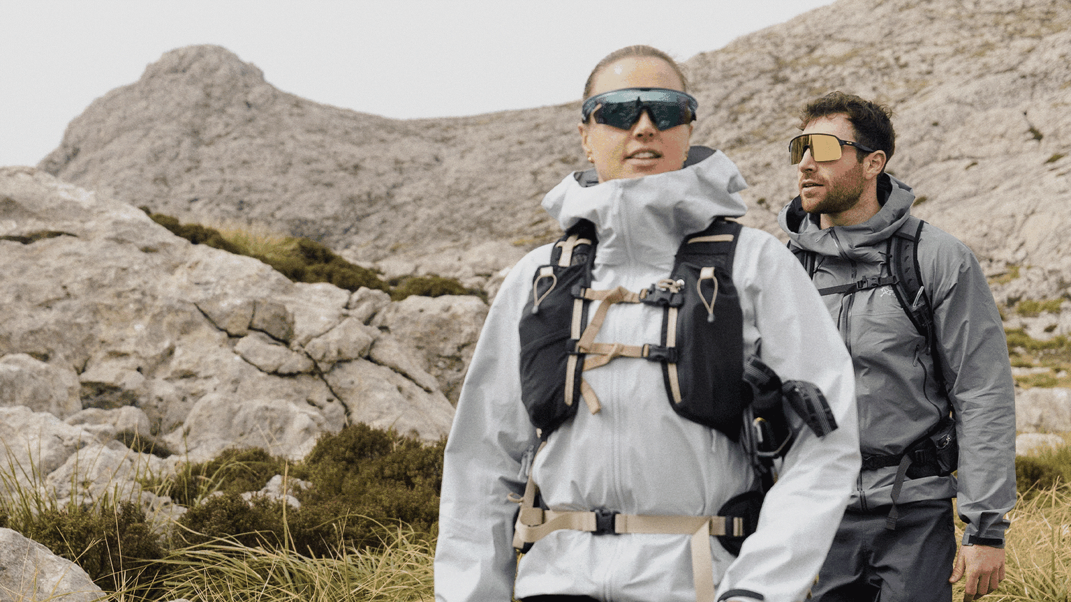 Two people in mountain gear hiking through rocky terrain, wearing sunglasses and jackets, with a backdrop of rugged peaks and grass.