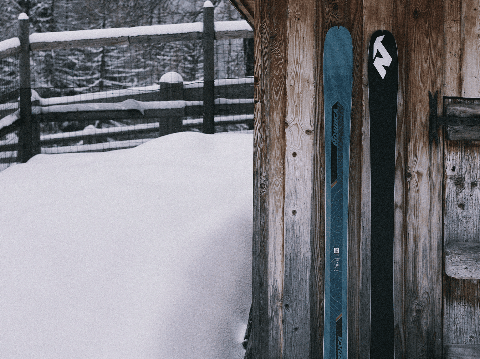 Two skis leaning against a wooden cabin, surrounded by snow and a rustic fence.