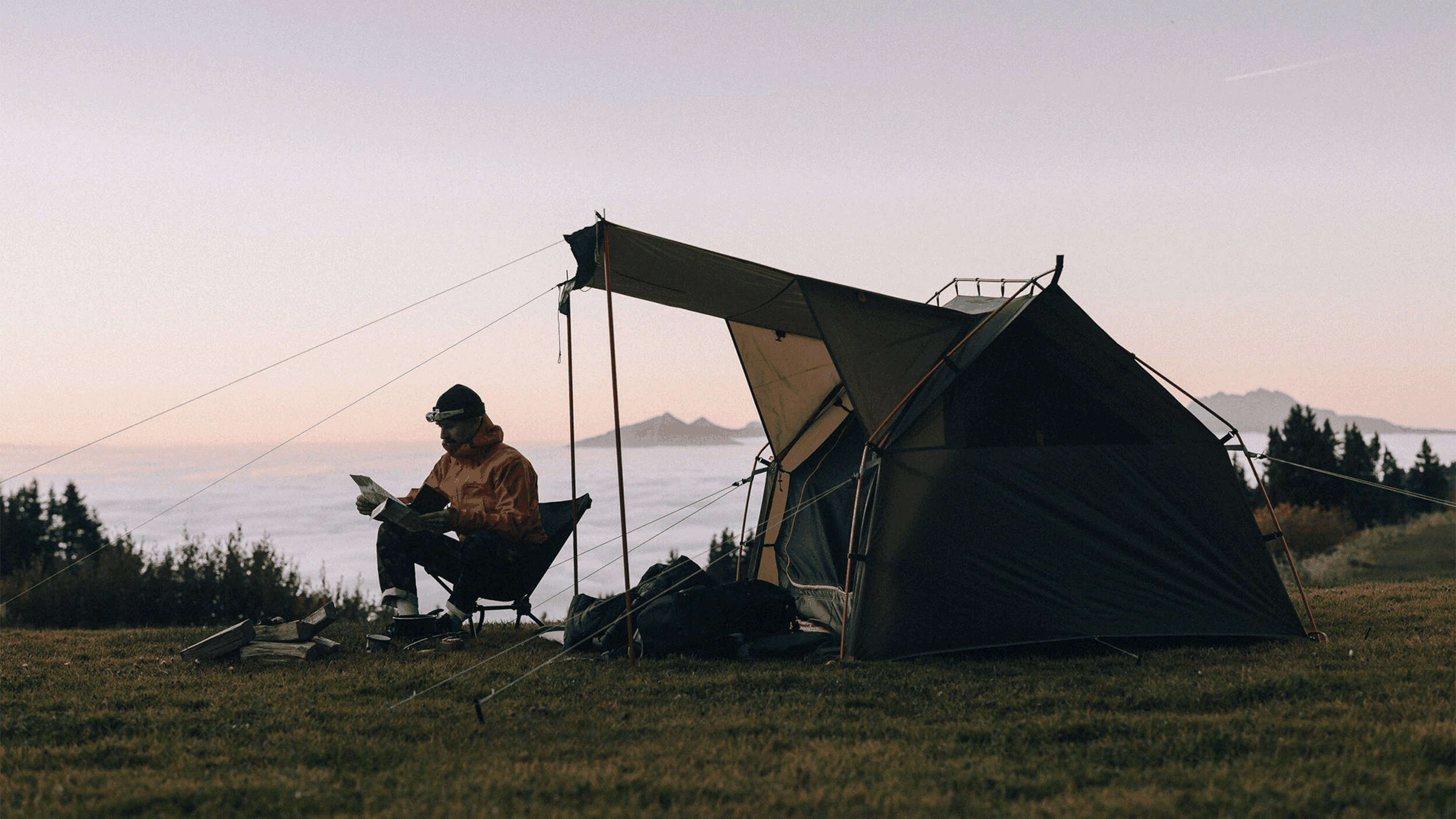 Camper in a red jacket reads beside a tent at sunrise, with mountains and a forest in the distance.