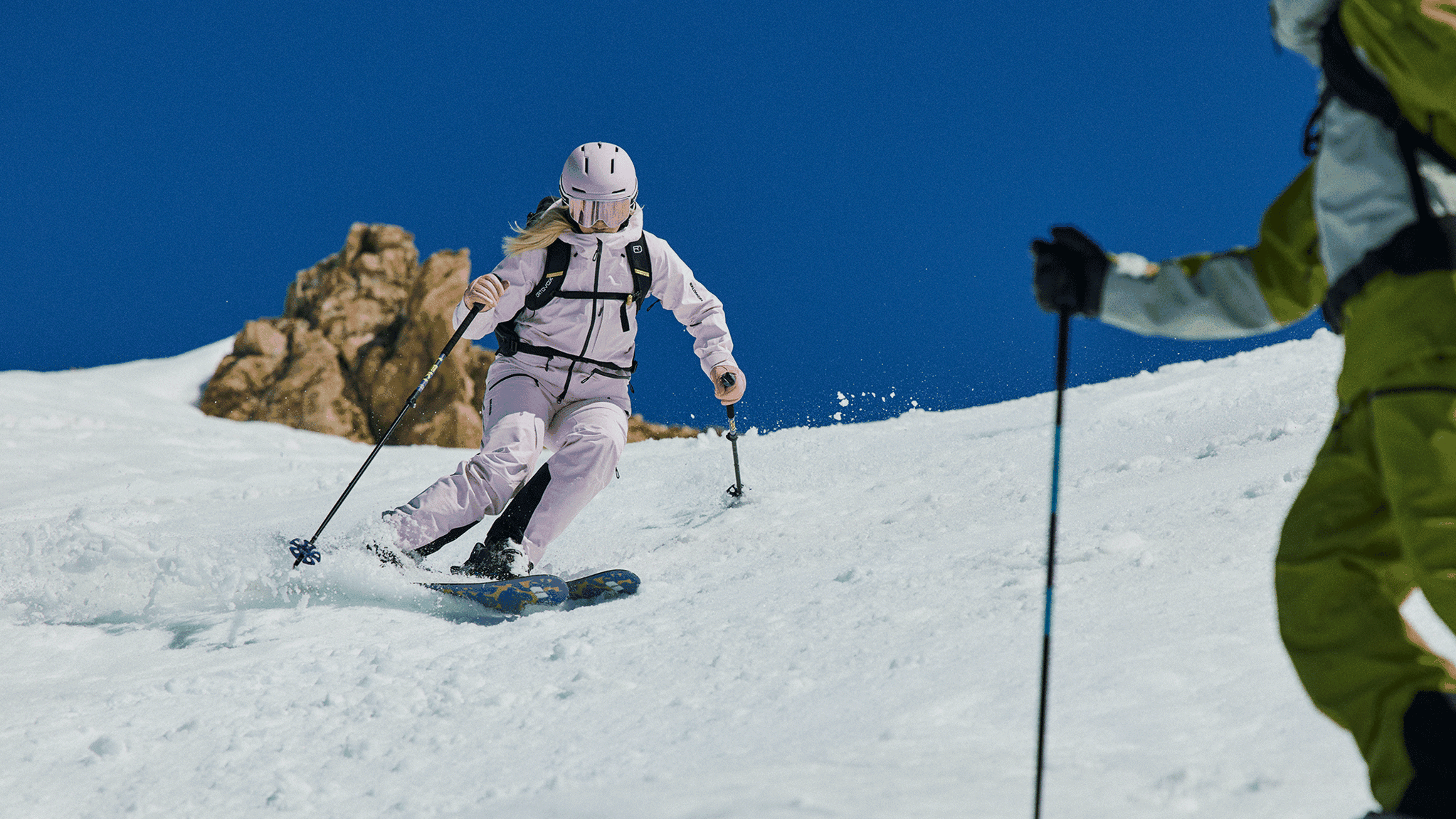 A skier in pink gear descends a snowy mountain slope under a clear blue sky, with rocky peaks in the background.