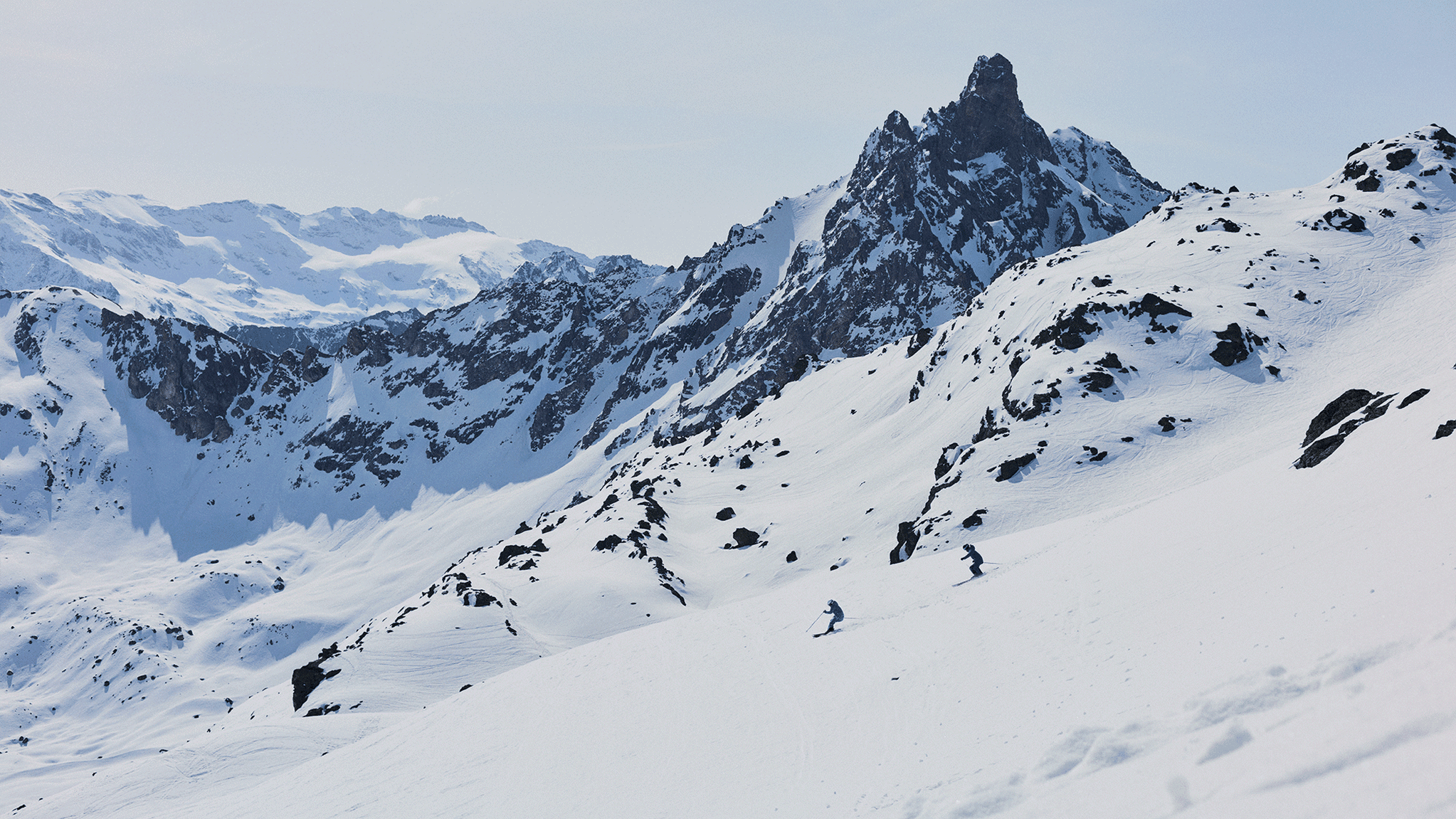 Two skiers dressed in blue descend a snowy mountain slope, surrounded by rugged snow-covered peaks under a clear sky.