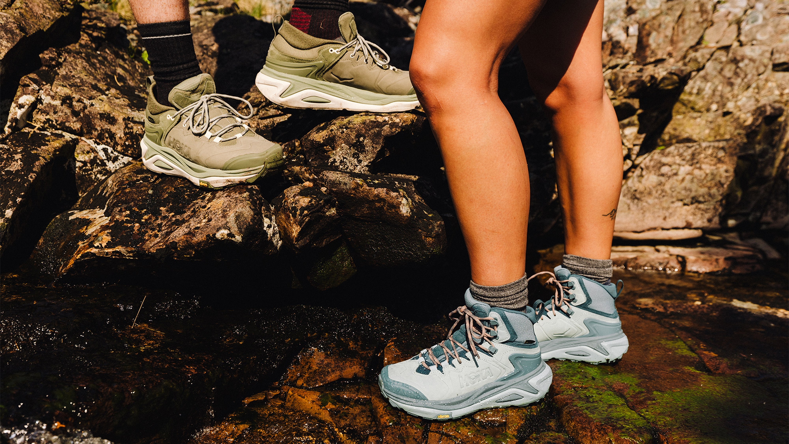 Two people wearing hiking boots stand on rocky terrain, one in olive green boots and the other in light blue.