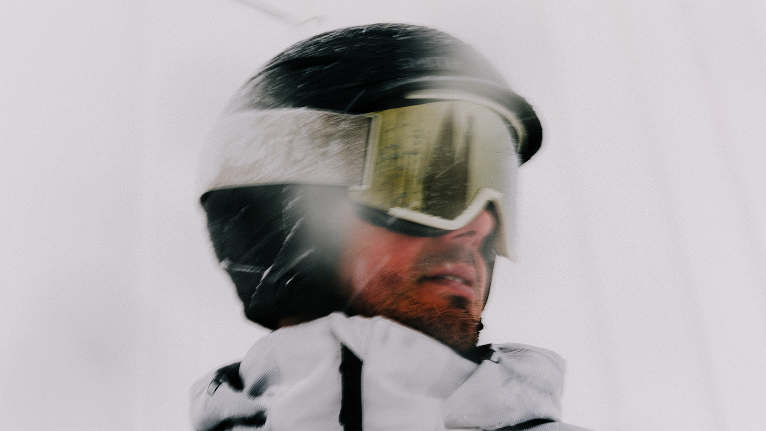 A person wearing a black helmet and reflective ski goggles, dressed in a winter jacket, with snow falling around them.