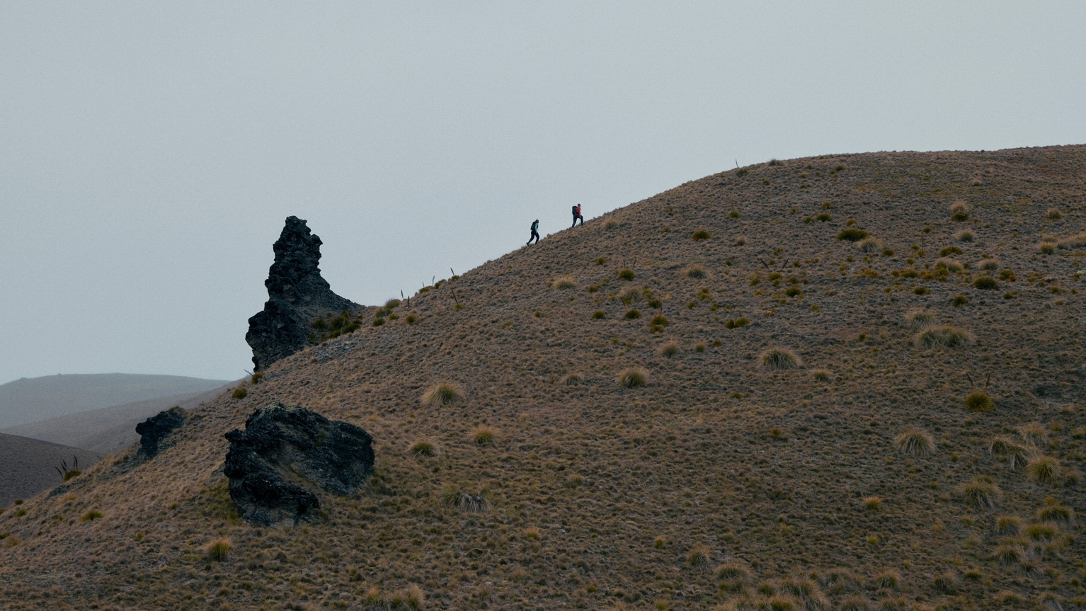 Two hikers ascend a grassy hill with scattered rocks under a grey sky, showcasing the rugged terrain and expansive landscape.
