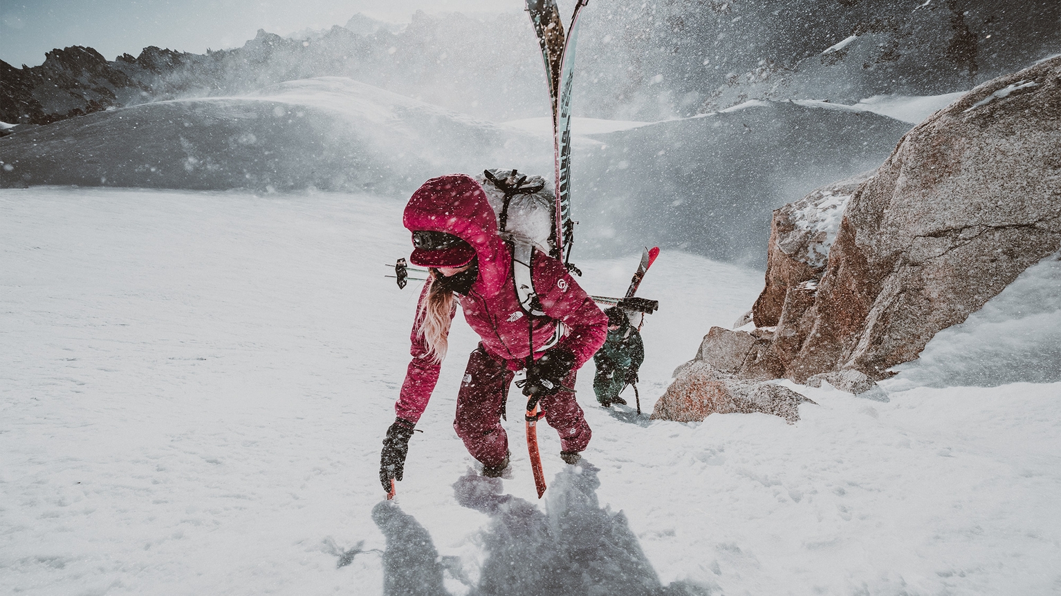 A person in red winter gear climbs a snowy mountain with skis on their back, using an ice axe for support, surrounded by falling snow.