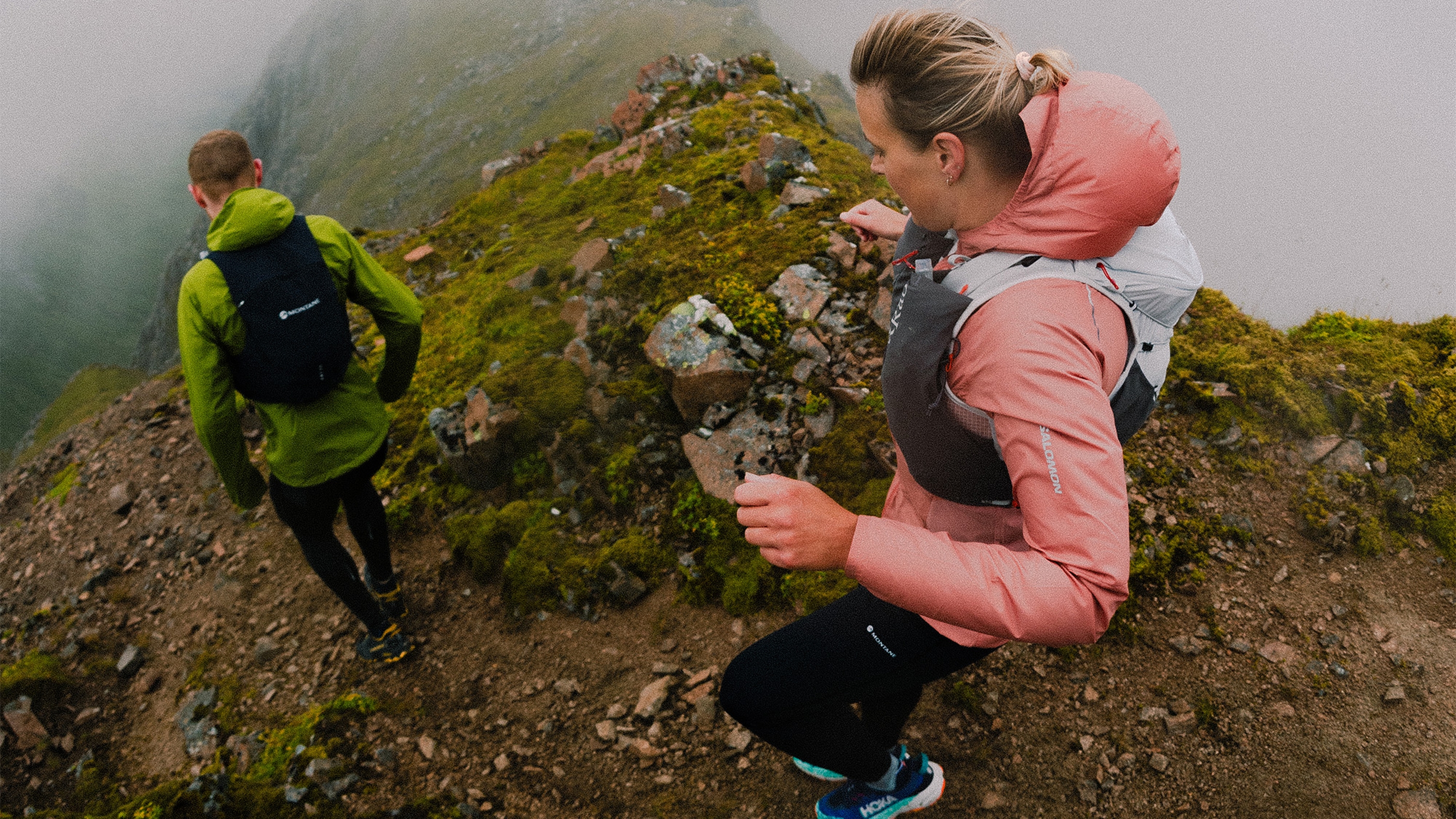 Two people trail running on a misty mountain path, wearing backpacks and colorful jackets, surrounded by green moss and rocky terrain.