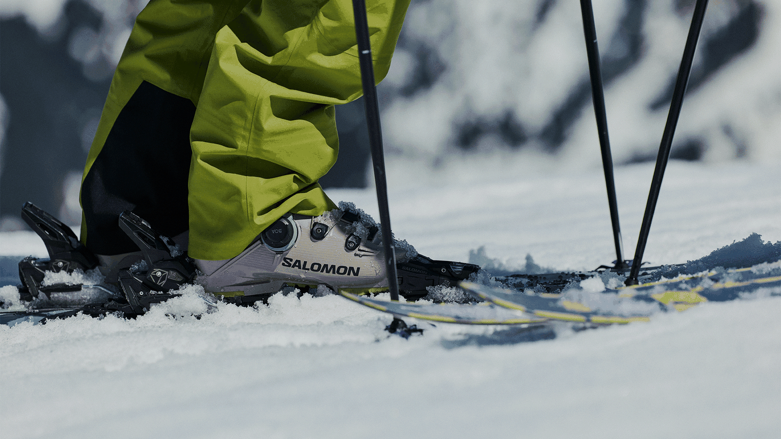 Skier wearing lime green pants and Salomon boots, preparing to ski on snow, with poles in hand.