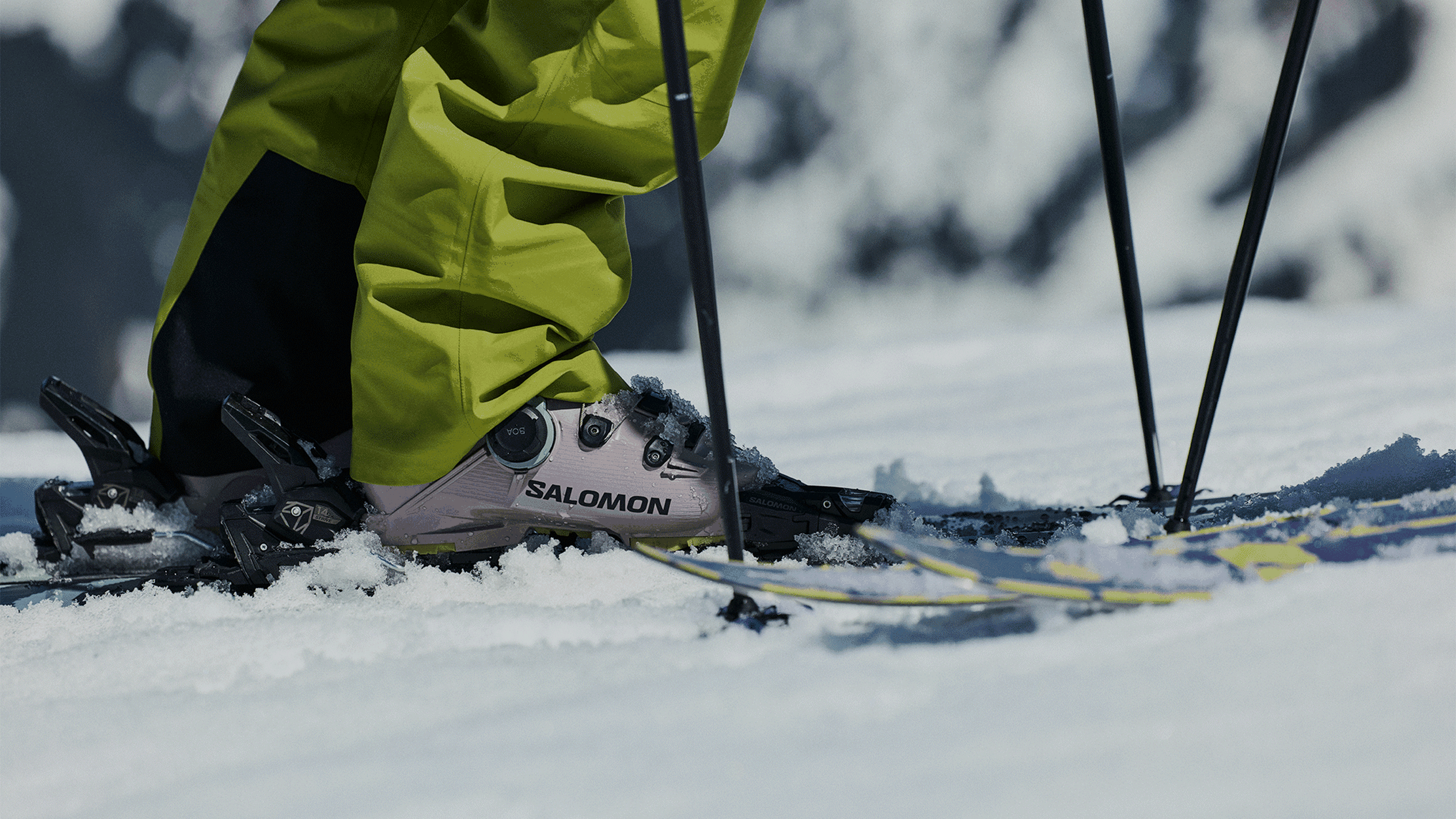 Skier wearing lime green pants and Salomon boots, preparing to ski on snow, with poles in hand.
