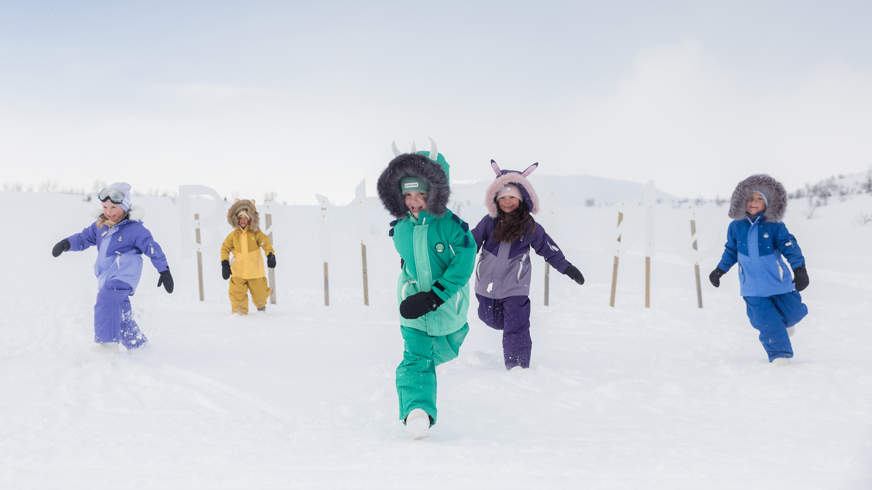 Five children in colourful insulated snowsuits and fur-trimmed hoods model mountain sports gear, striding through deep snow on an overcast plain.