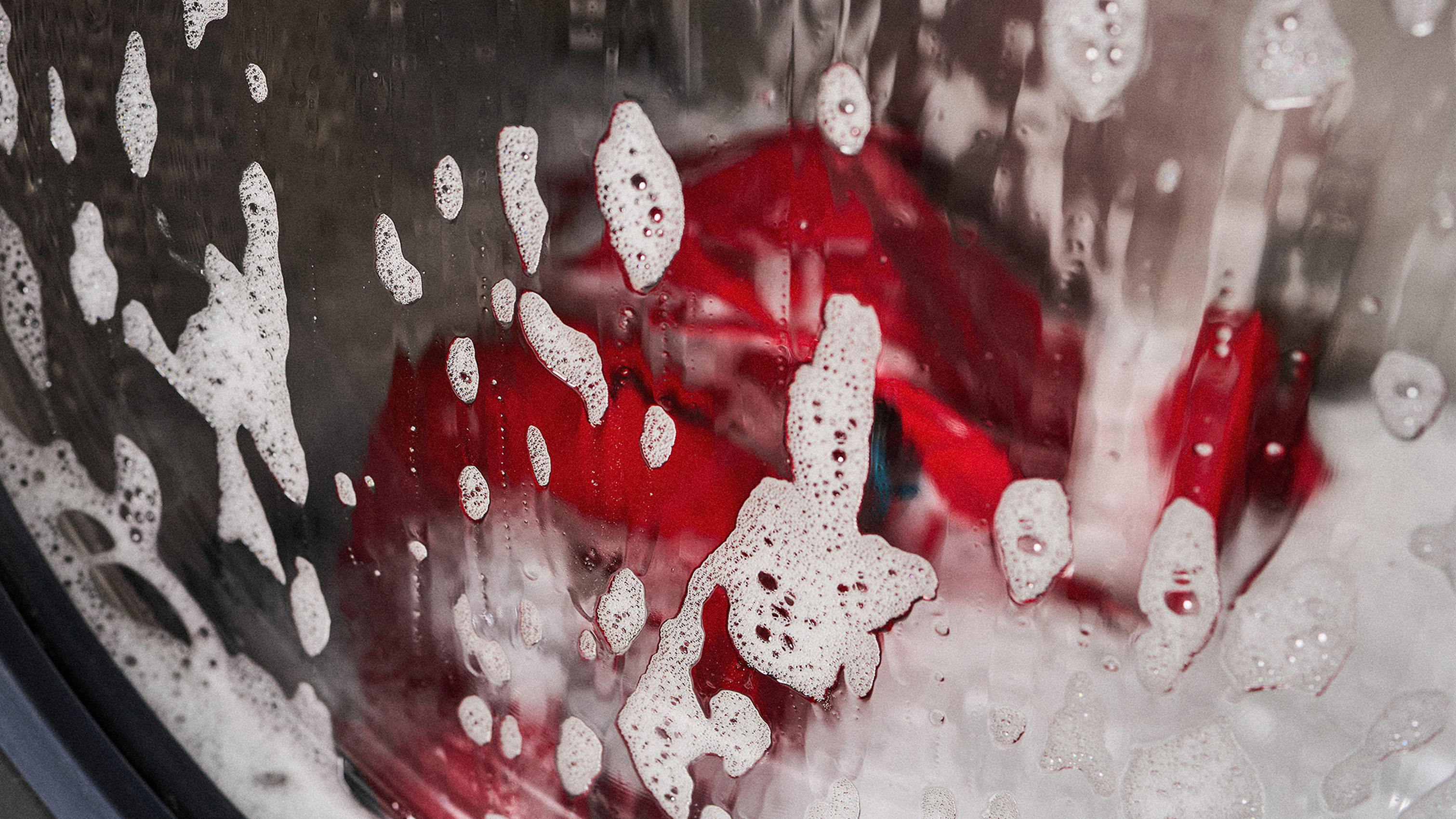 Soap suds on a washing machine window with red clothing visible inside during a wash cycle.