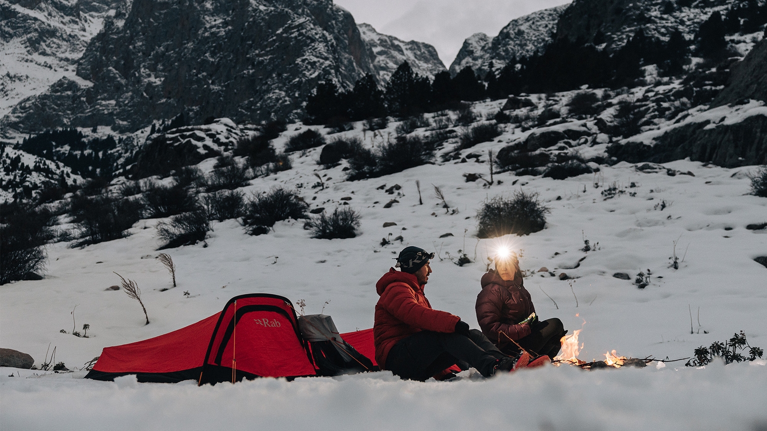 Two people sit by a campfire in snowy mountains, next to a red bivvy bag, wearing winter gear and headlamps, surrounded by rugged peaks.