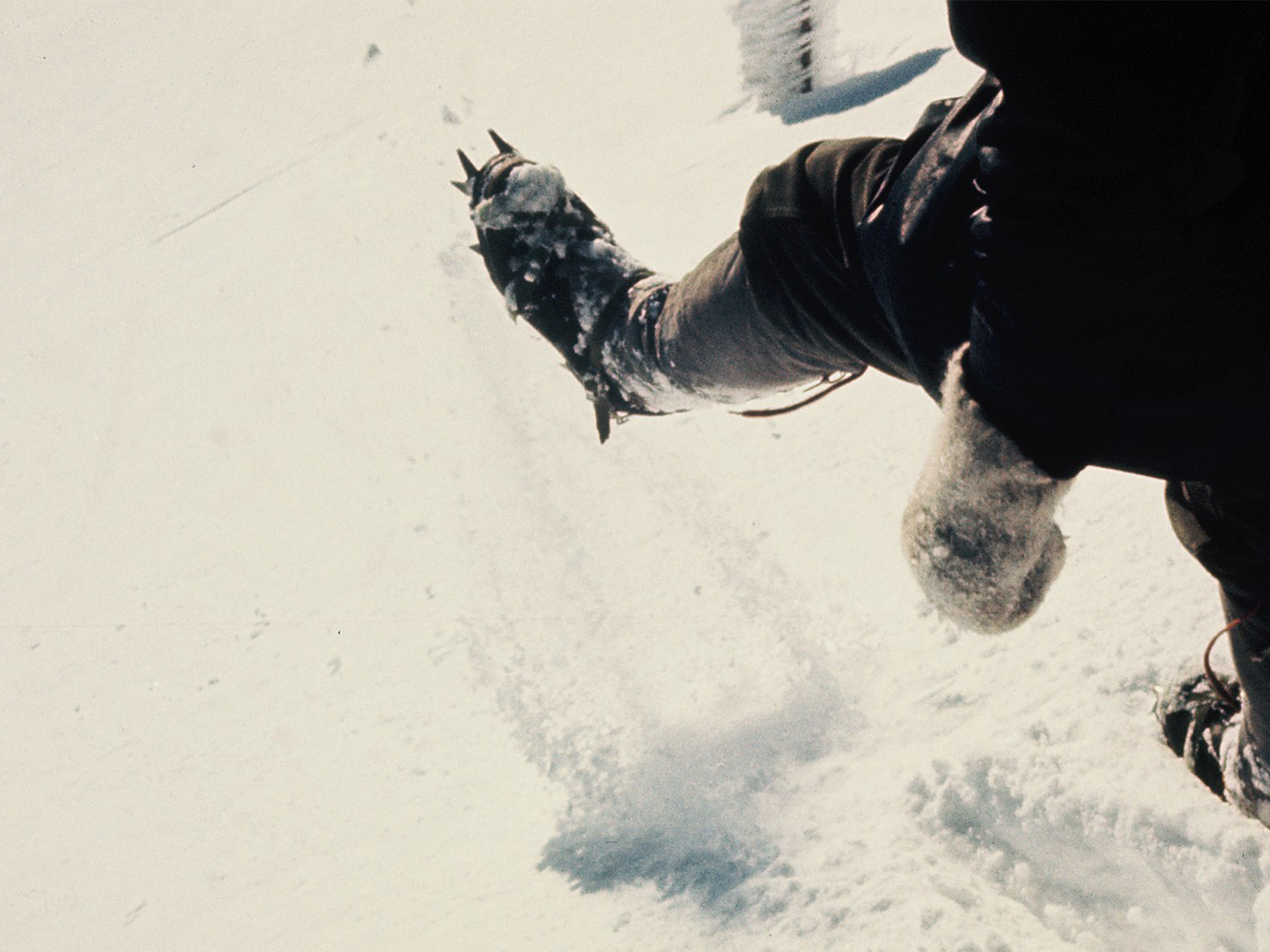 Close-up of a person wearing crampons on snowy terrain, kicking up snow as they climb or hike.