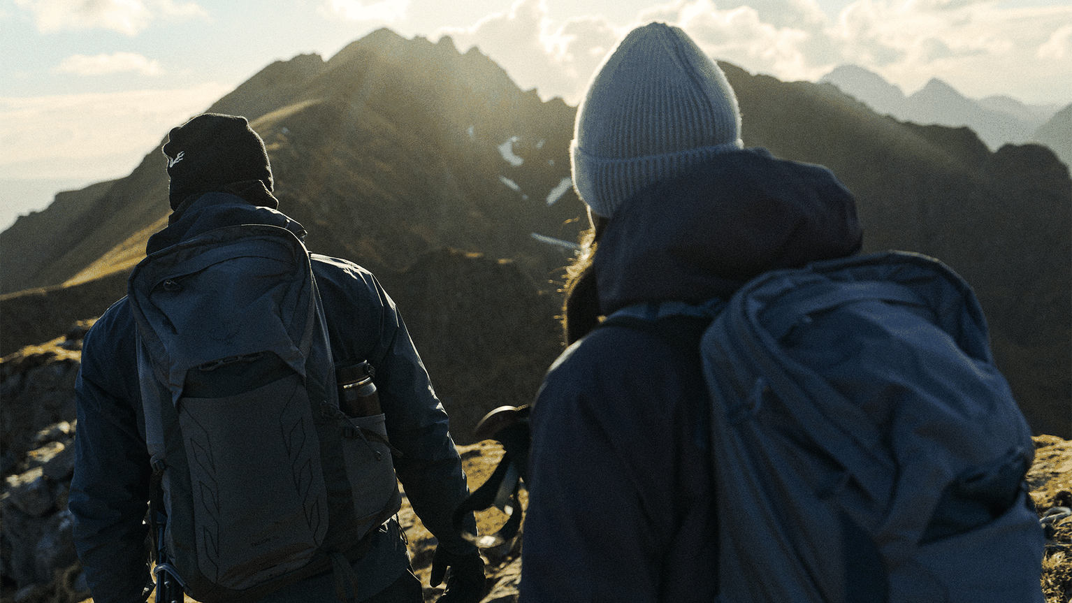Two hikers with backpacks trek a mountain ridge at sunrise, with rugged peaks in the background.