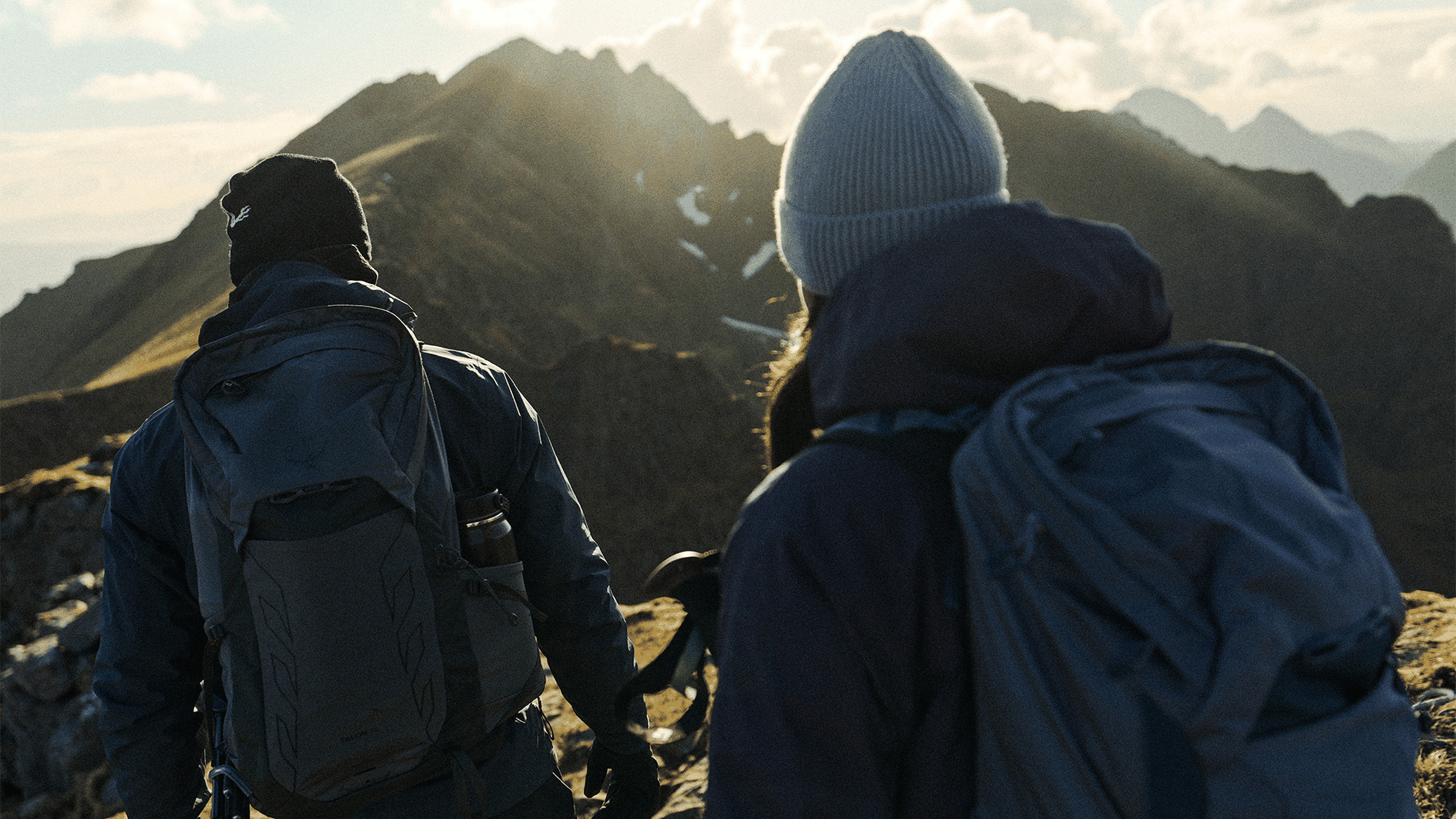 Two hikers with backpacks trek a mountain ridge at sunrise, with rugged peaks in the background.