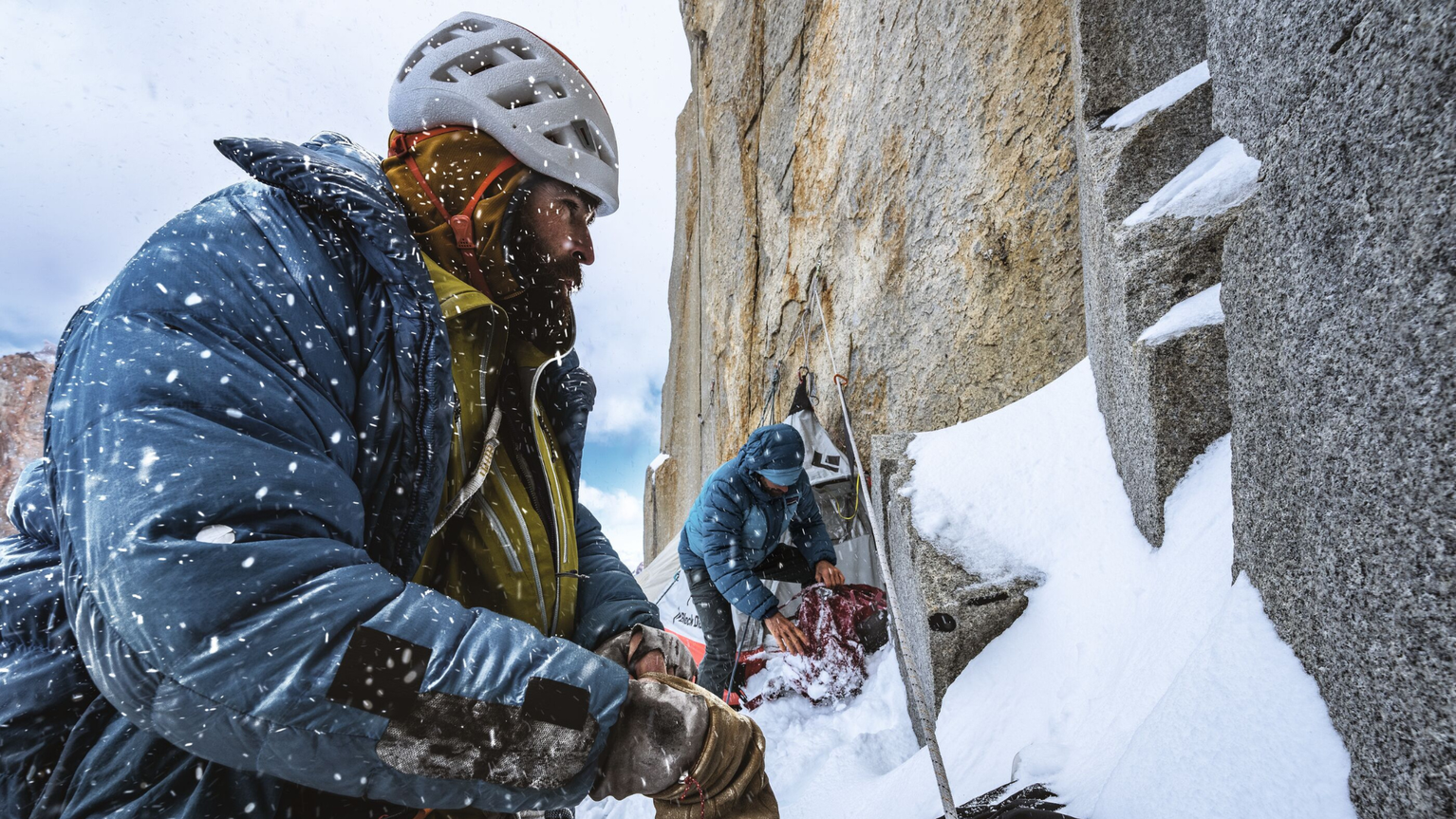 Climbers in winter gear ascend a snowy, rocky mountain face, focused and determined amidst falling snowflakes.