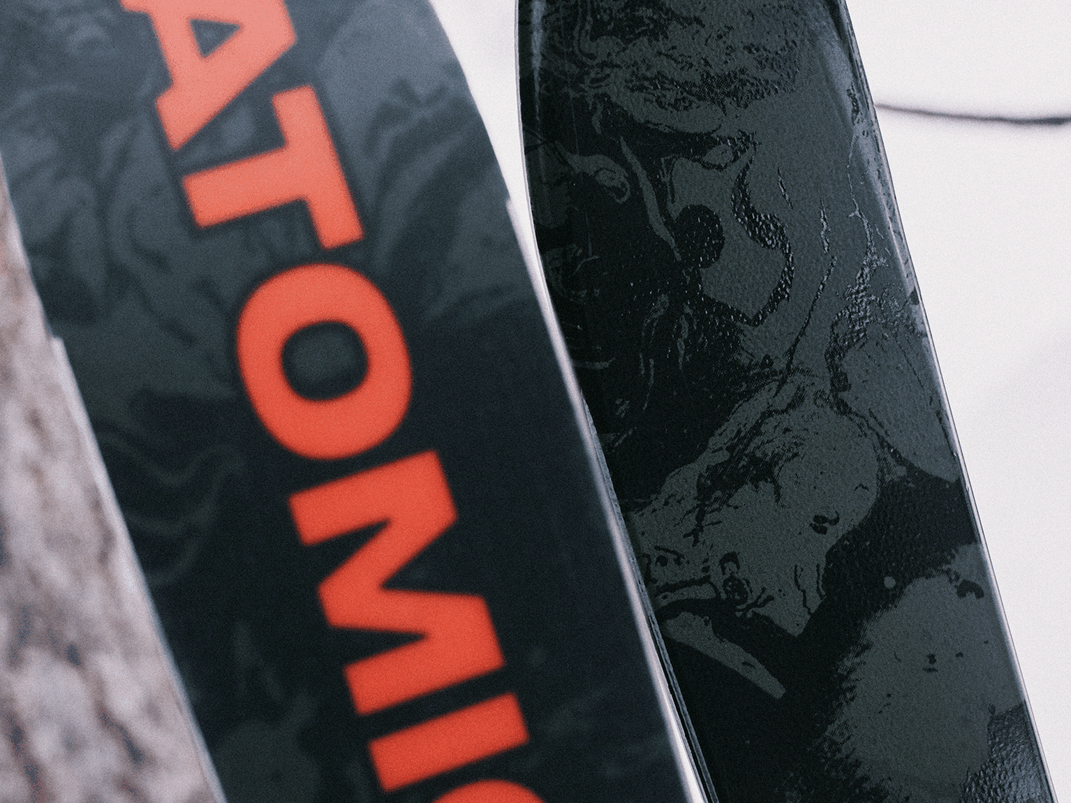 Close-up of two skis, one with a black and grey pattern and the other featuring the red word "Atomic" against a snowy background.