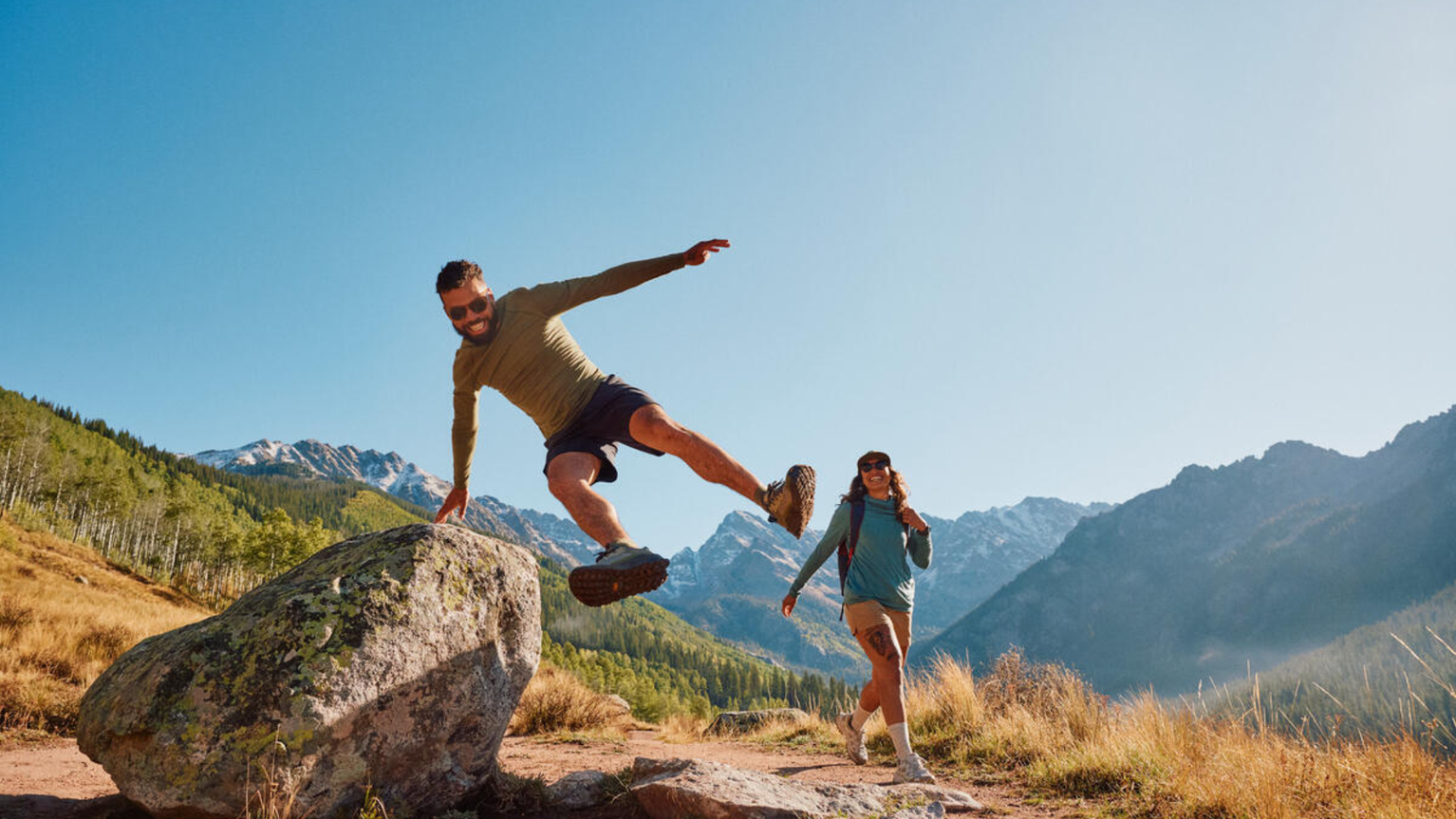 Man and woman enjoying a hike in a mountainous area, with the man playfully leaping over a rock under a clear blue sky.