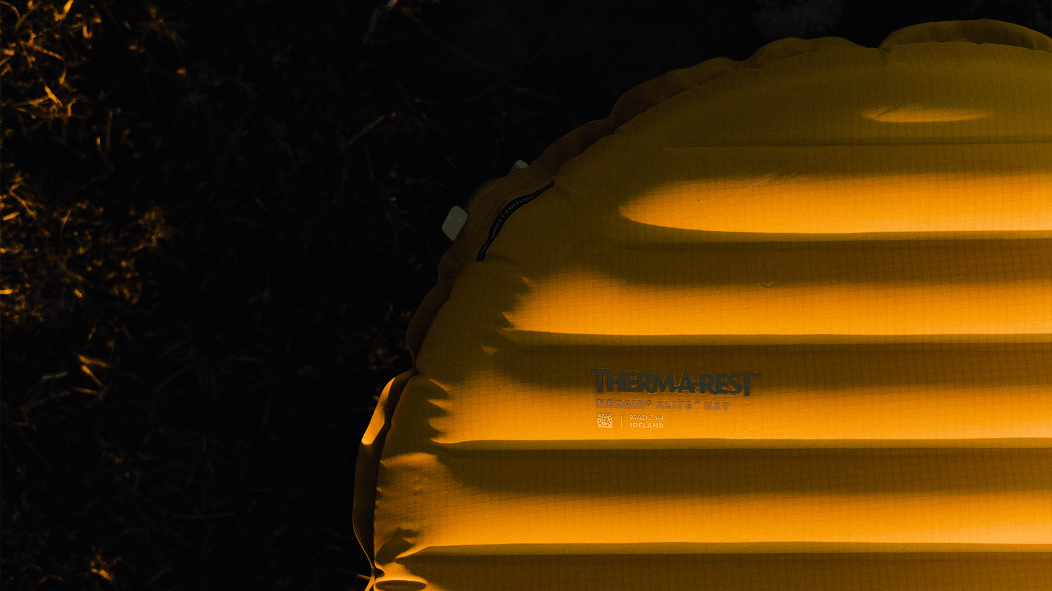 Yellow inflatable camping mat, partially visible, laying on dark ground, with branding text on the surface.