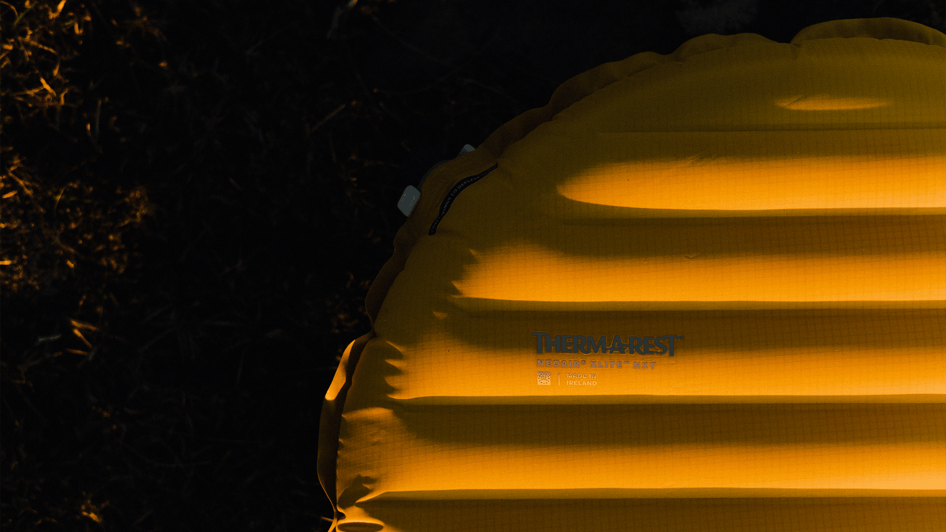 Yellow inflatable camping mat, partially visible, laying on dark ground, with branding text on the surface.