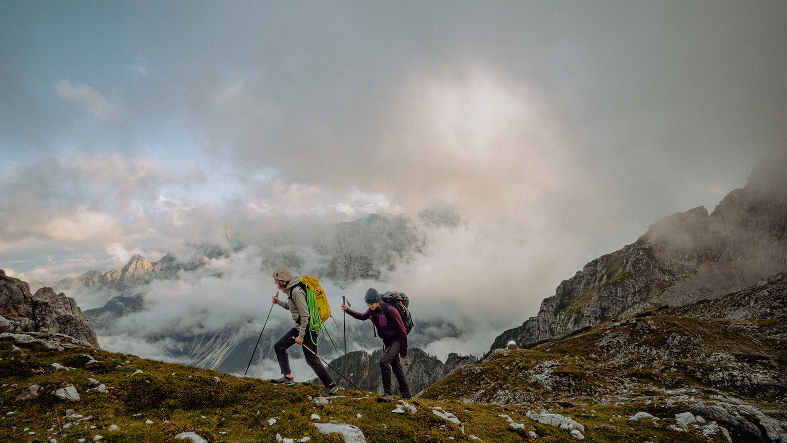 Two hikers with backpacks and trekking poles traverse a rugged mountain trail under a cloudy sky.