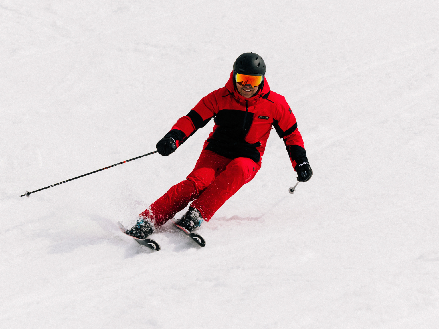Skier in red technical kit carves a clean turn on packed snow, wearing helmet and mirrored goggles.