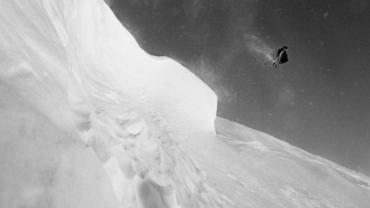A skier performs a high jump off a snowy slope against a clear sky in a black and white image.