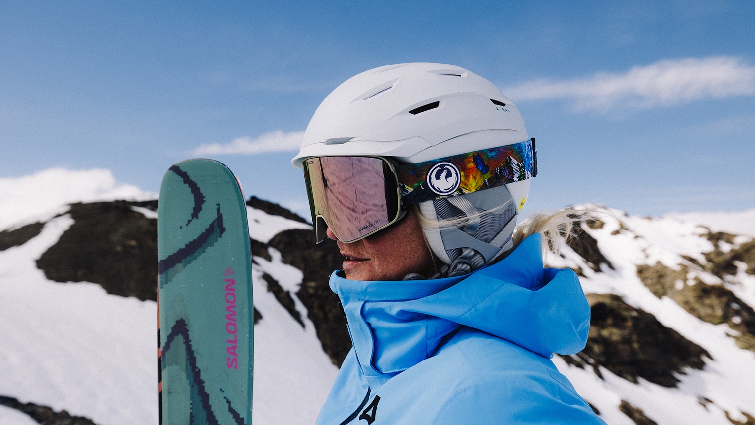 Person in blue jacket and white helmet holding skis, standing on a snowy mountain with a clear blue sky.