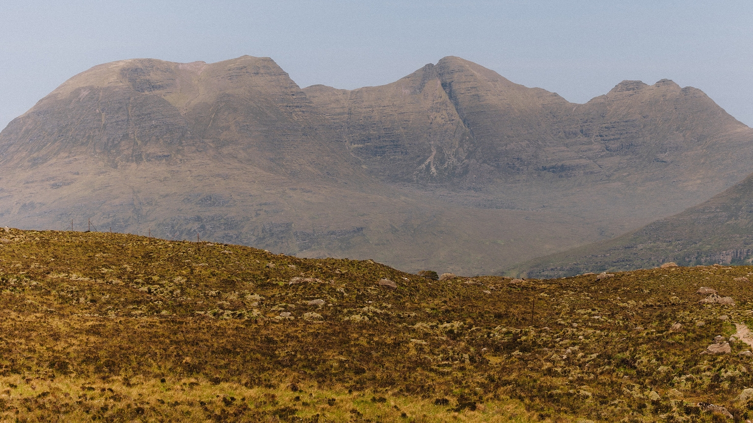 Distant view of rocky, rugged mountains under a clear blue sky, with a foreground of brownish grassland and scattered rocks.