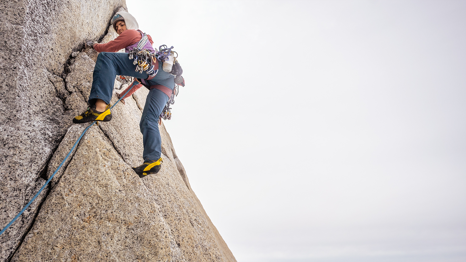 Person rock climbing on a steep rock face, wearing climbing gear, against a cloudy sky.
