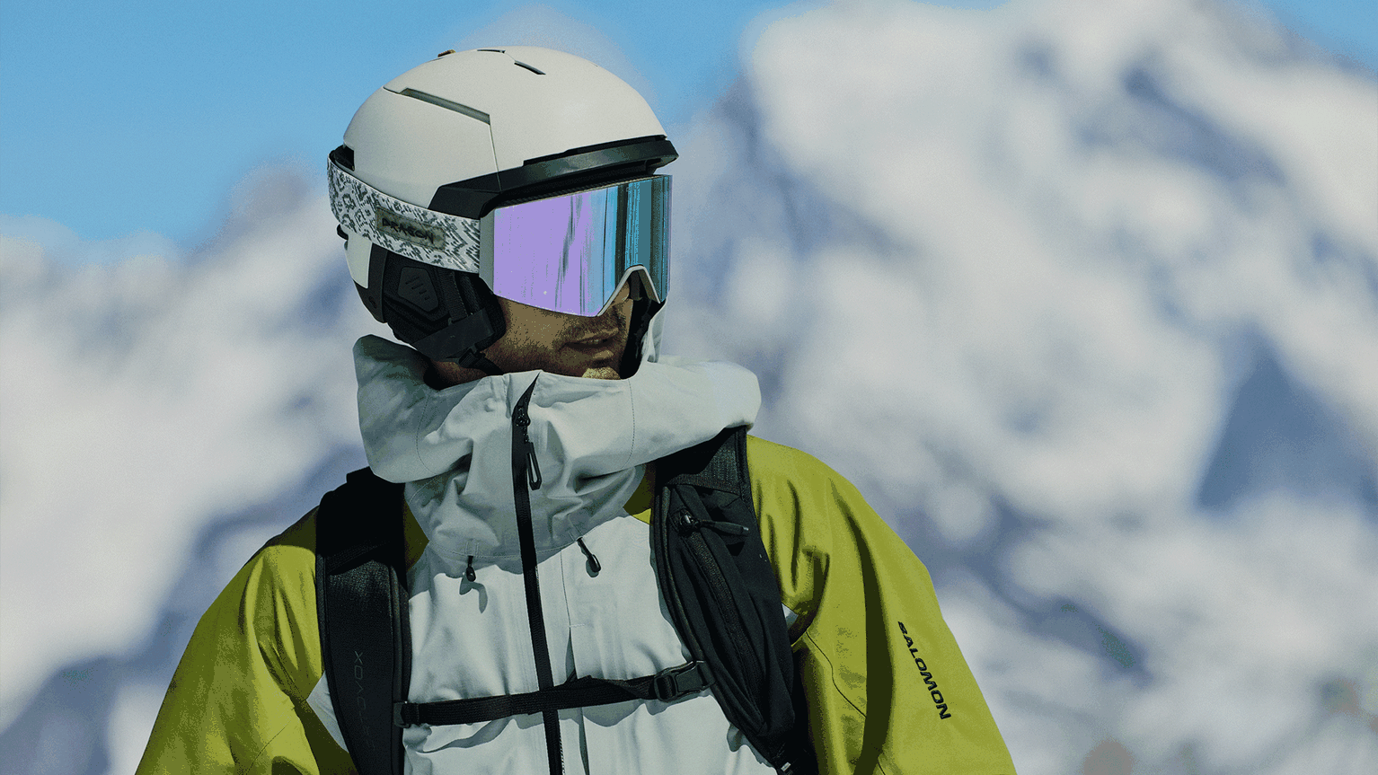 Man in a helmet and ski goggles wearing a green and white jacket, with snowy mountains in the background.