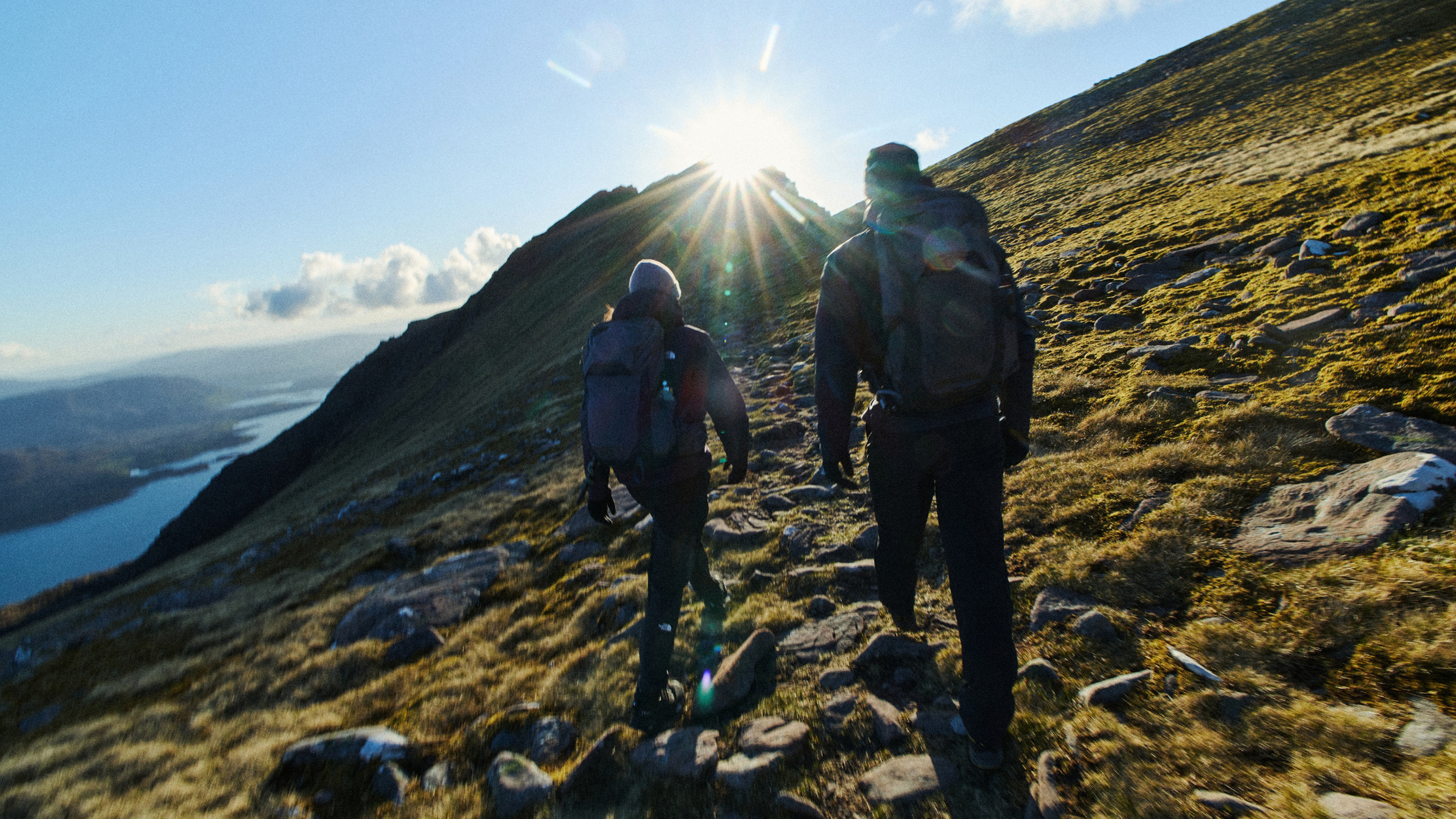 Two people with Osprey backpacks hiking up a sunlit mountain trail, surrounded by grassy terrain and a distant view of a lake.