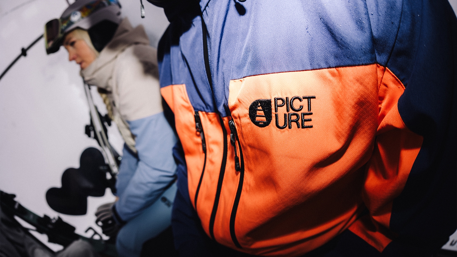 Close-up of a person wearing a blue and orange jacket with "Picture" logo. Another person with ski gear is in the background.